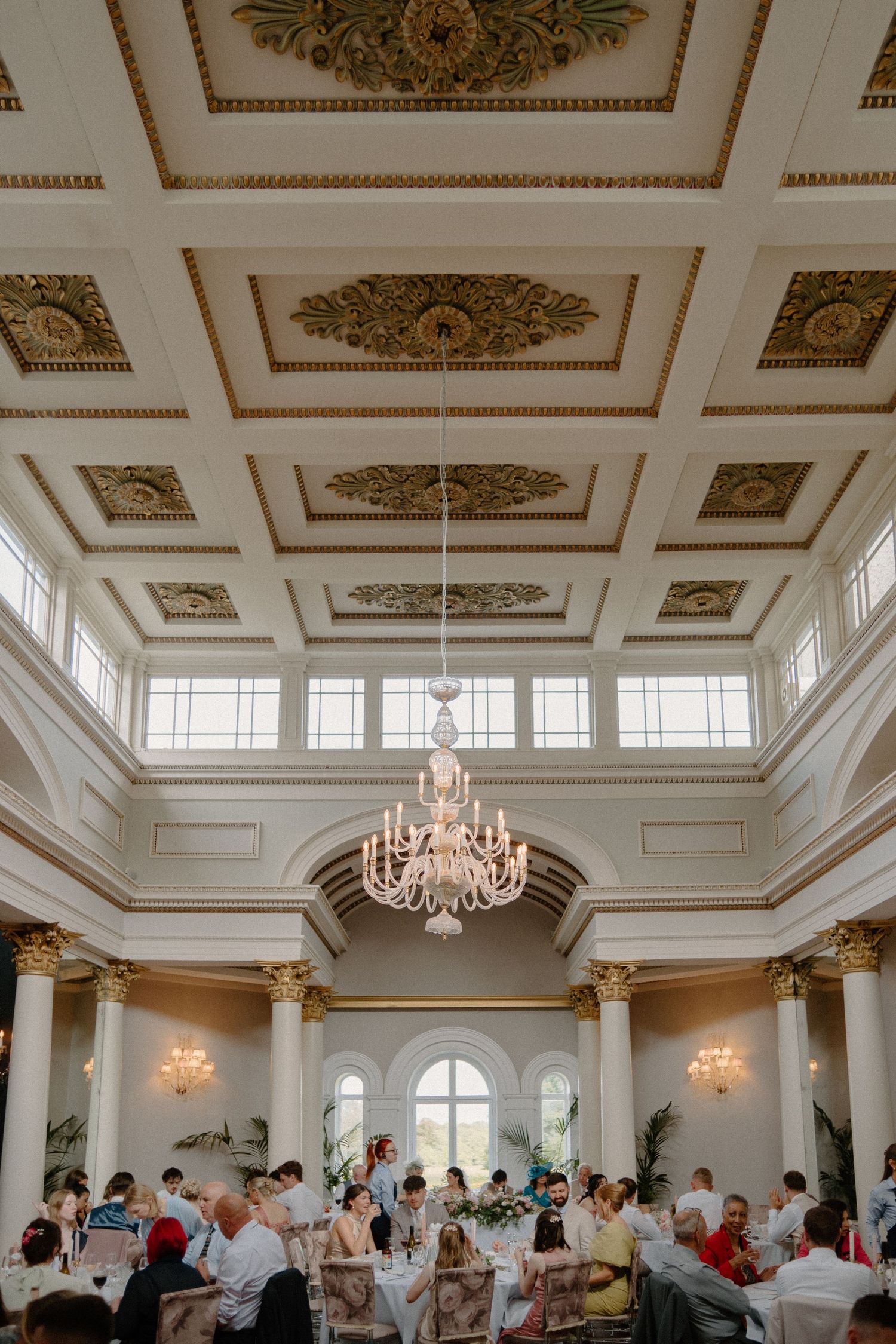 Grand wedding reception hall with ornate coffered ceiling, crystal chandelier, tall columns, and guests seated at tables.