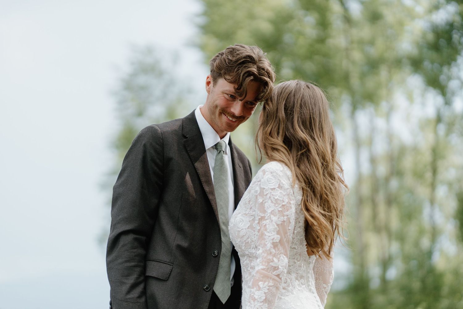 Bride and groom share an intimate smiling moment together outdoors among tall green trees.