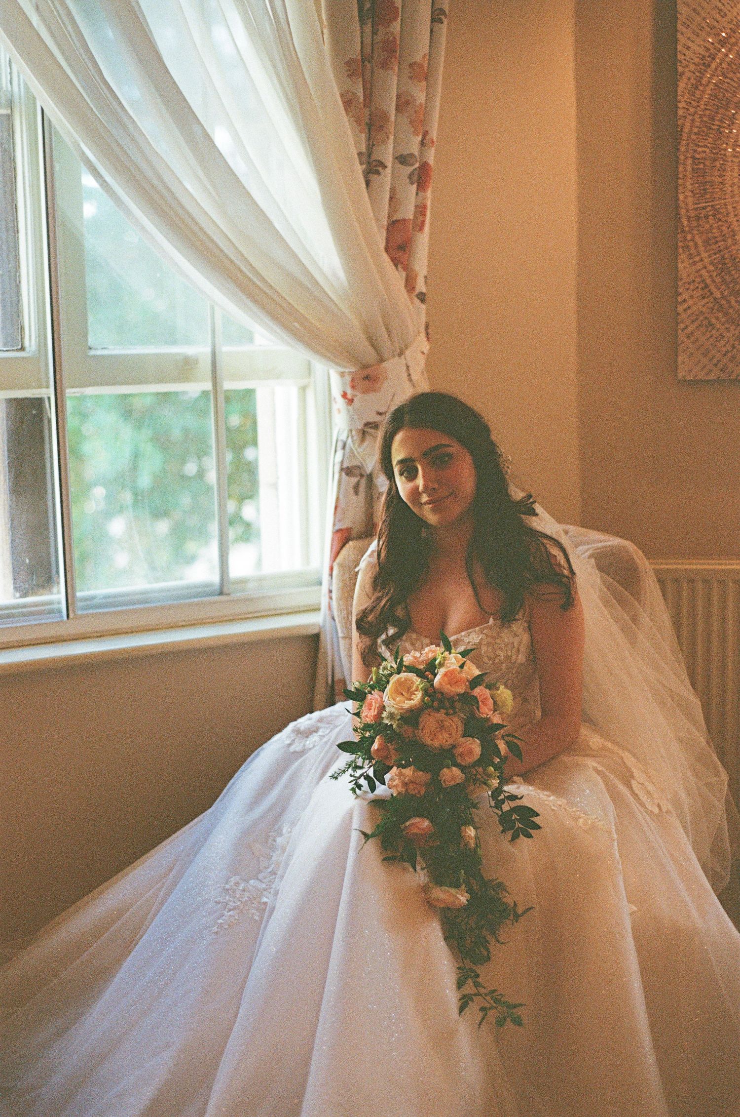 Bride in ball gown with pink bouquet seated by a sunlit window with sheer curtains.