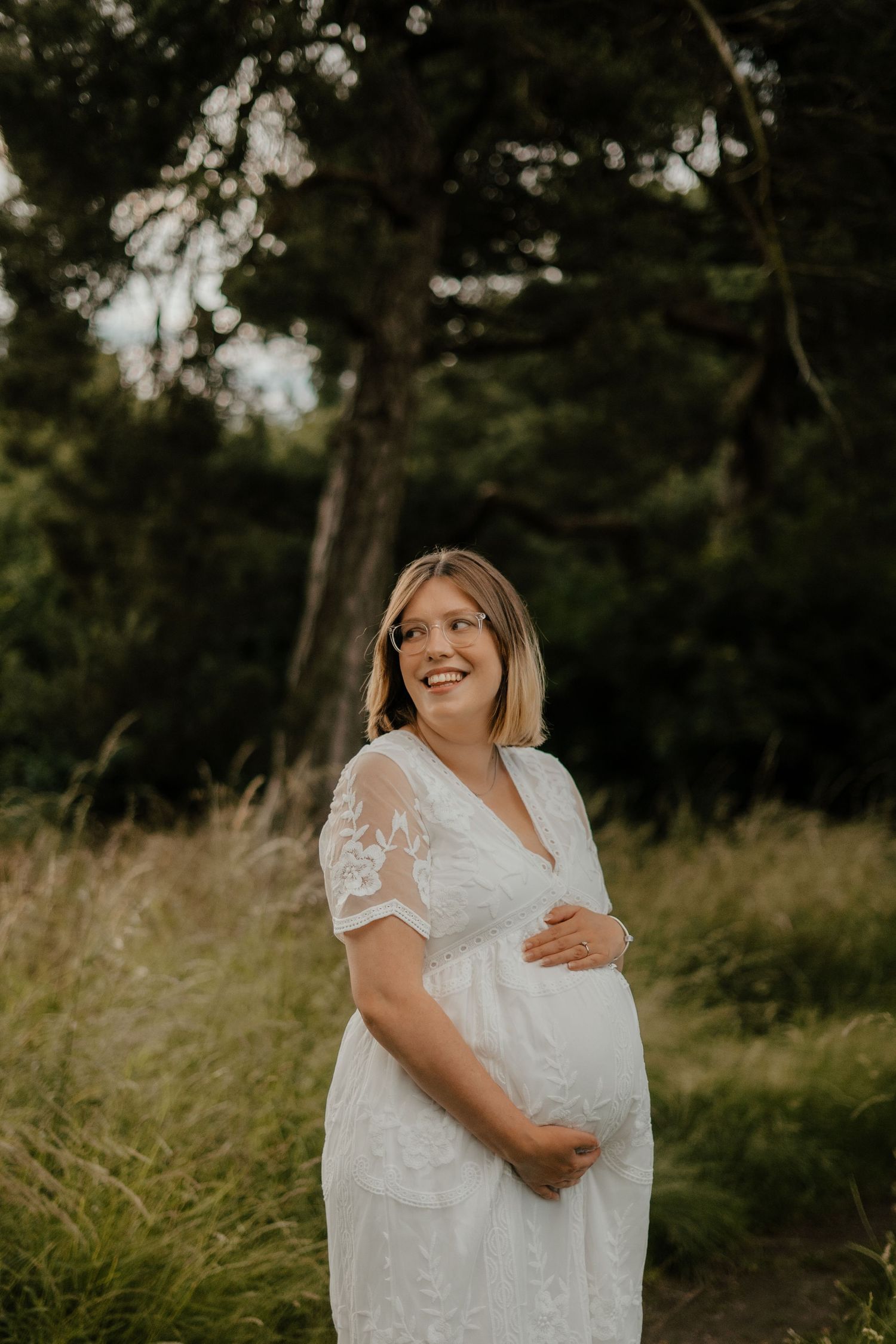 A smiling pregnant woman in a white lace dress cradles her bump in a forest setting.