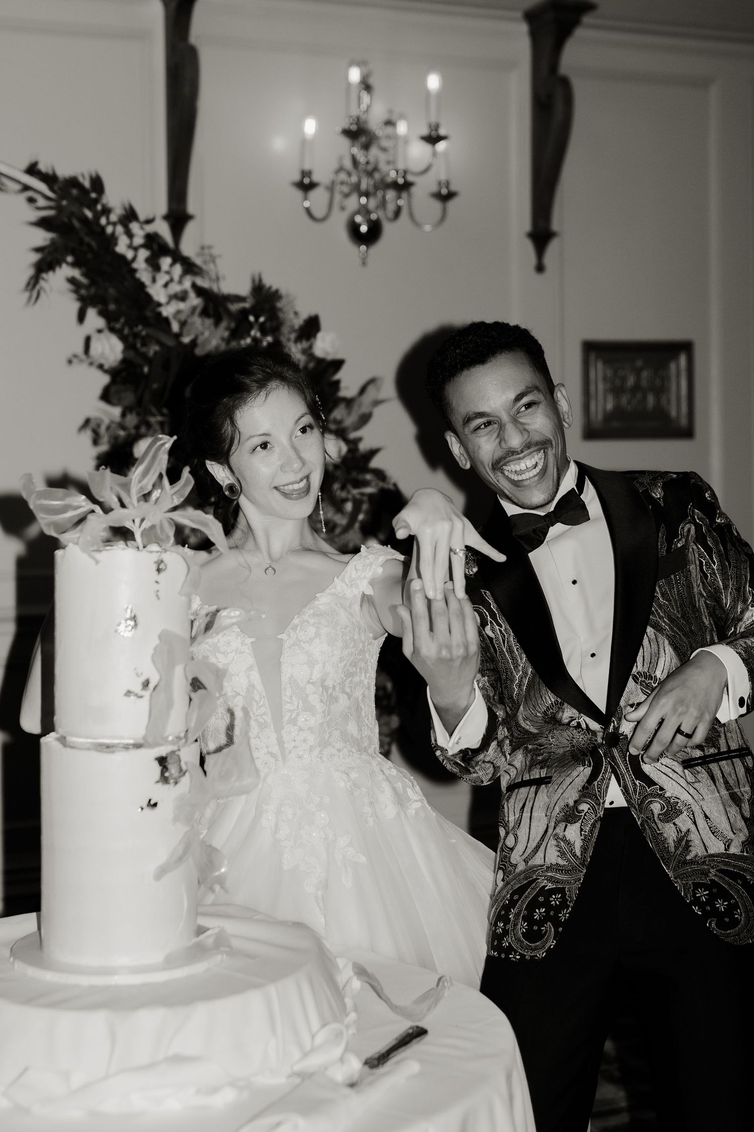Black and white photo of laughing newlyweds cutting tiered wedding cake, groom in ornate gold blazer.