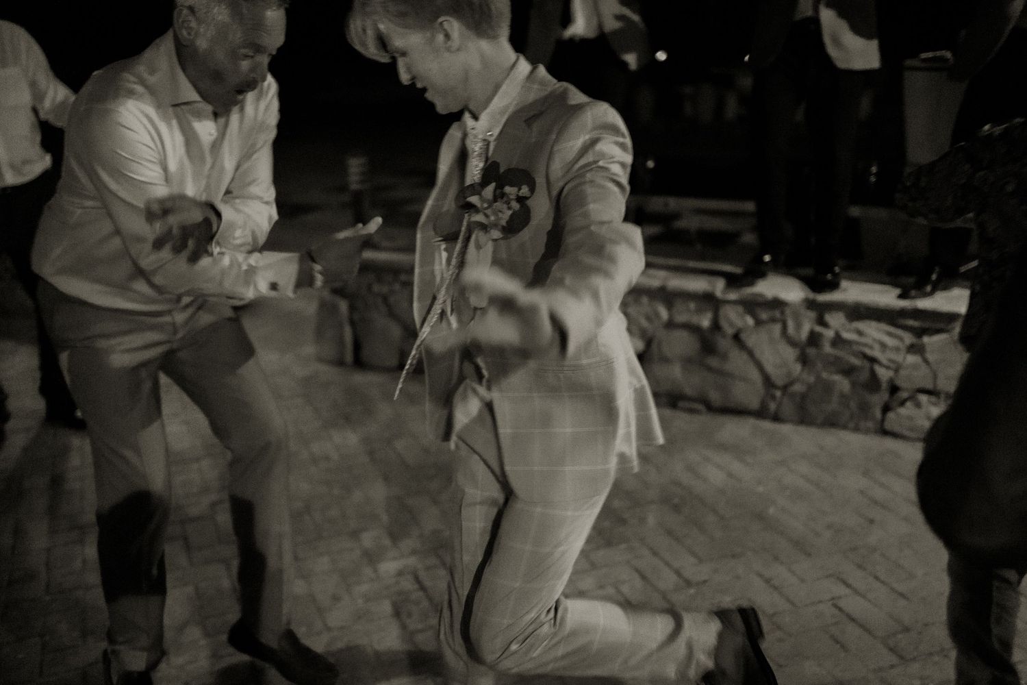Black and white photo of groom on one knee dancing at outdoor wedding reception.