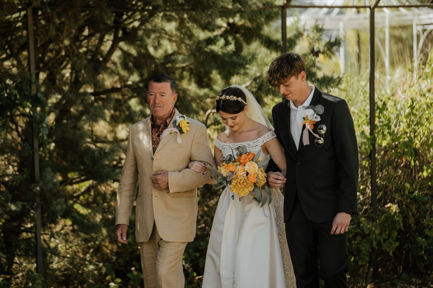 Bride in white off-shoulder gown walks down the aisle escorted by two men in a sunlit garden setting.