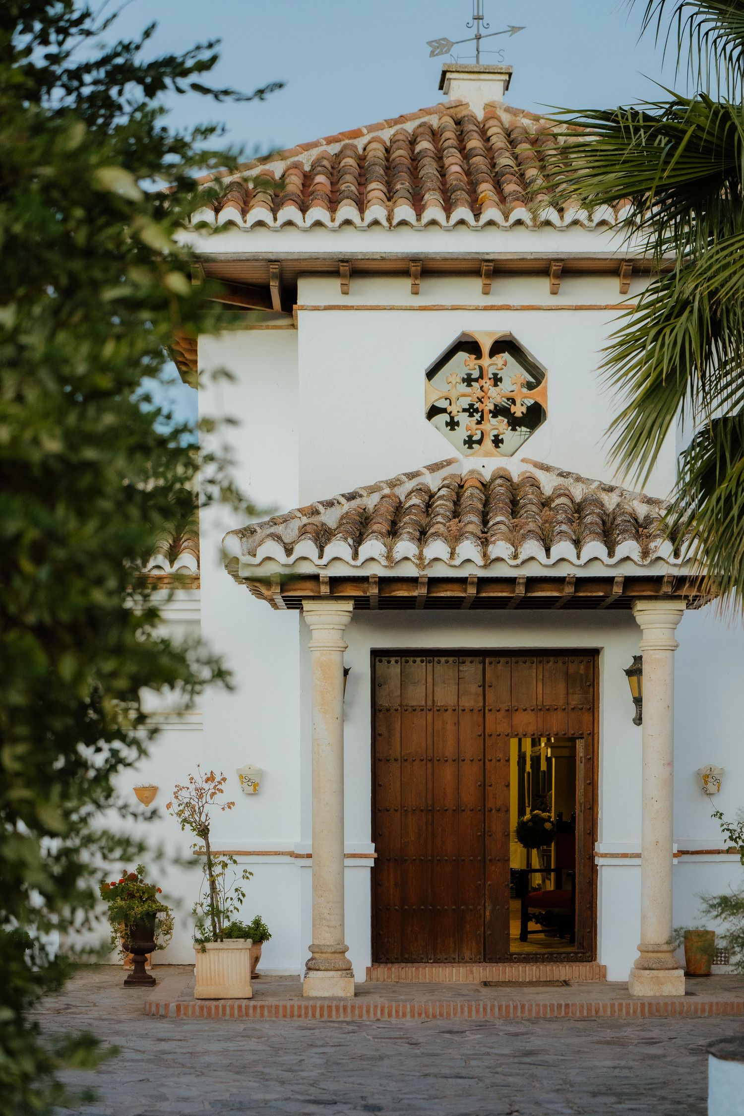 White Spanish chapel with terracotta roof tiles and wooden doors framed by palm trees.