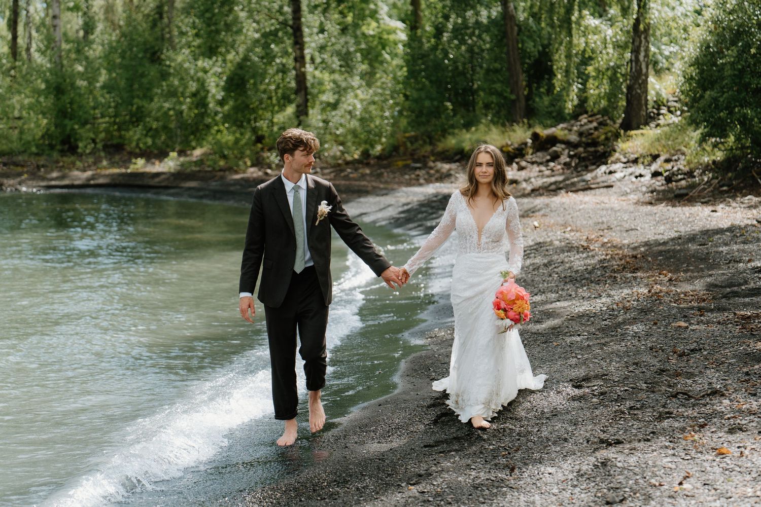 Bride and groom walking hand-in-hand along a river's edge in a forest setting.