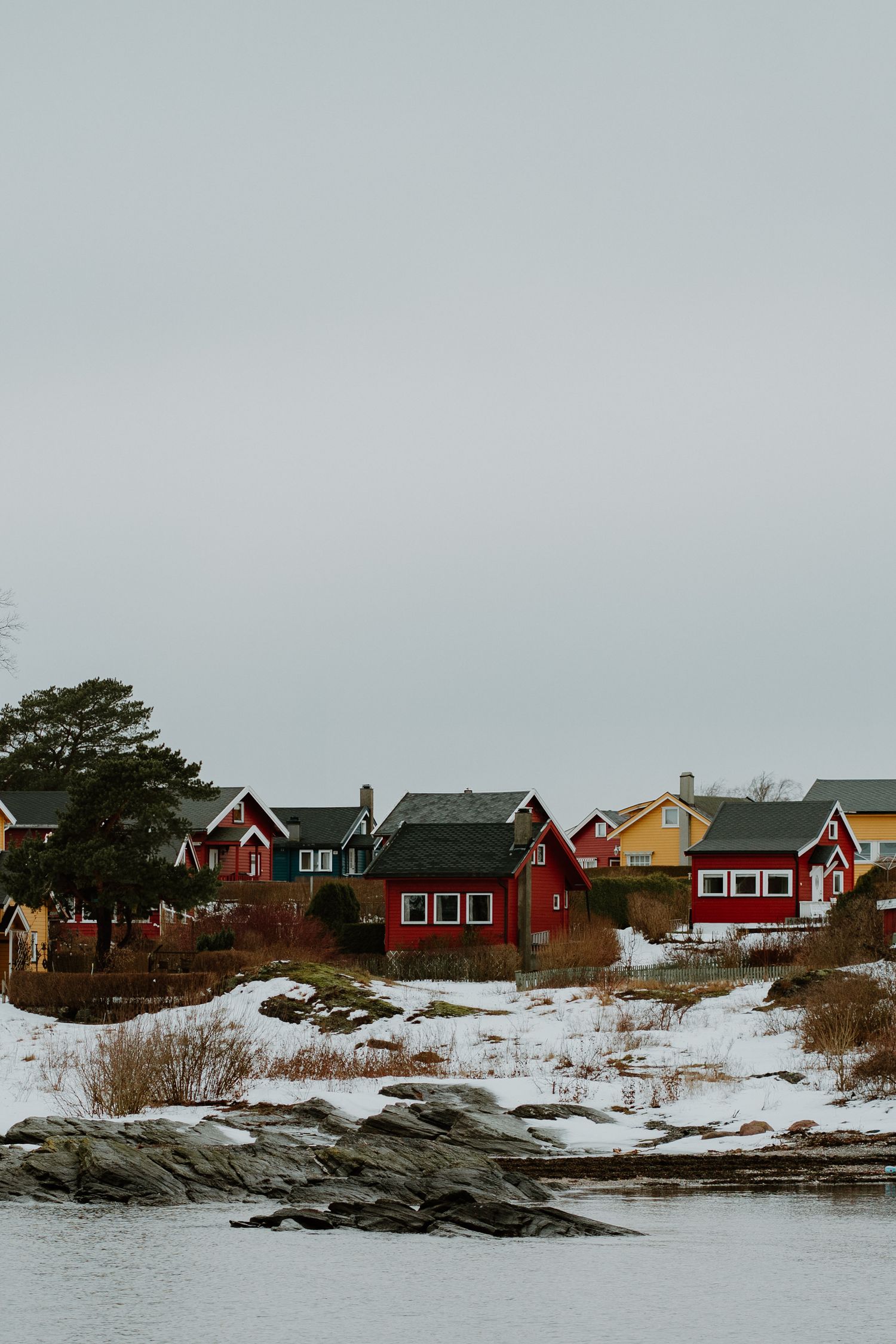 Red and yellow coastal houses dot a snowy shoreline with rocky outcrops along a gray winter waterfront in Scandinavia.