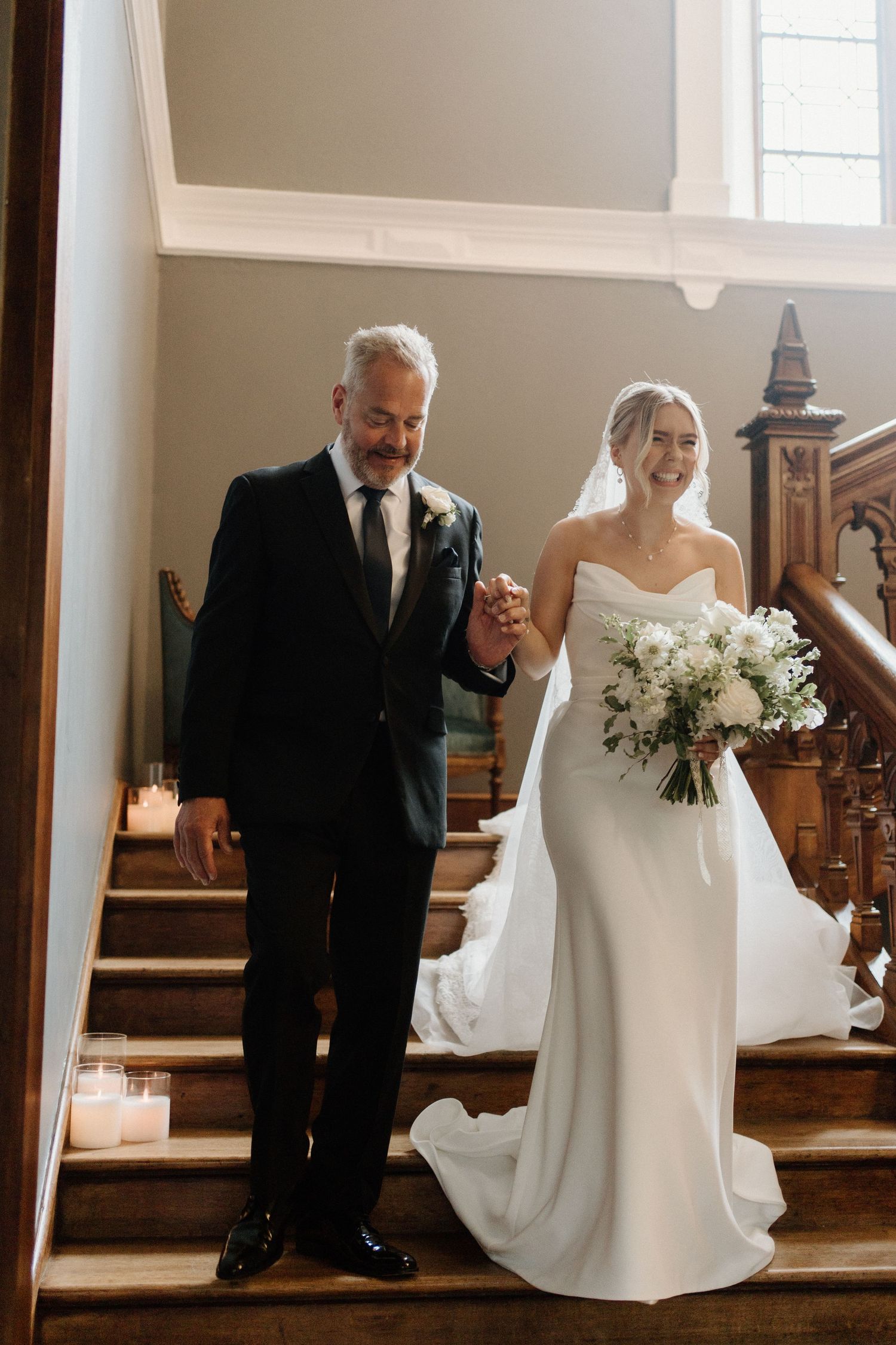 Bride in white gown holds bouquet while descending candlelit staircase with older gentleman in dark suit.