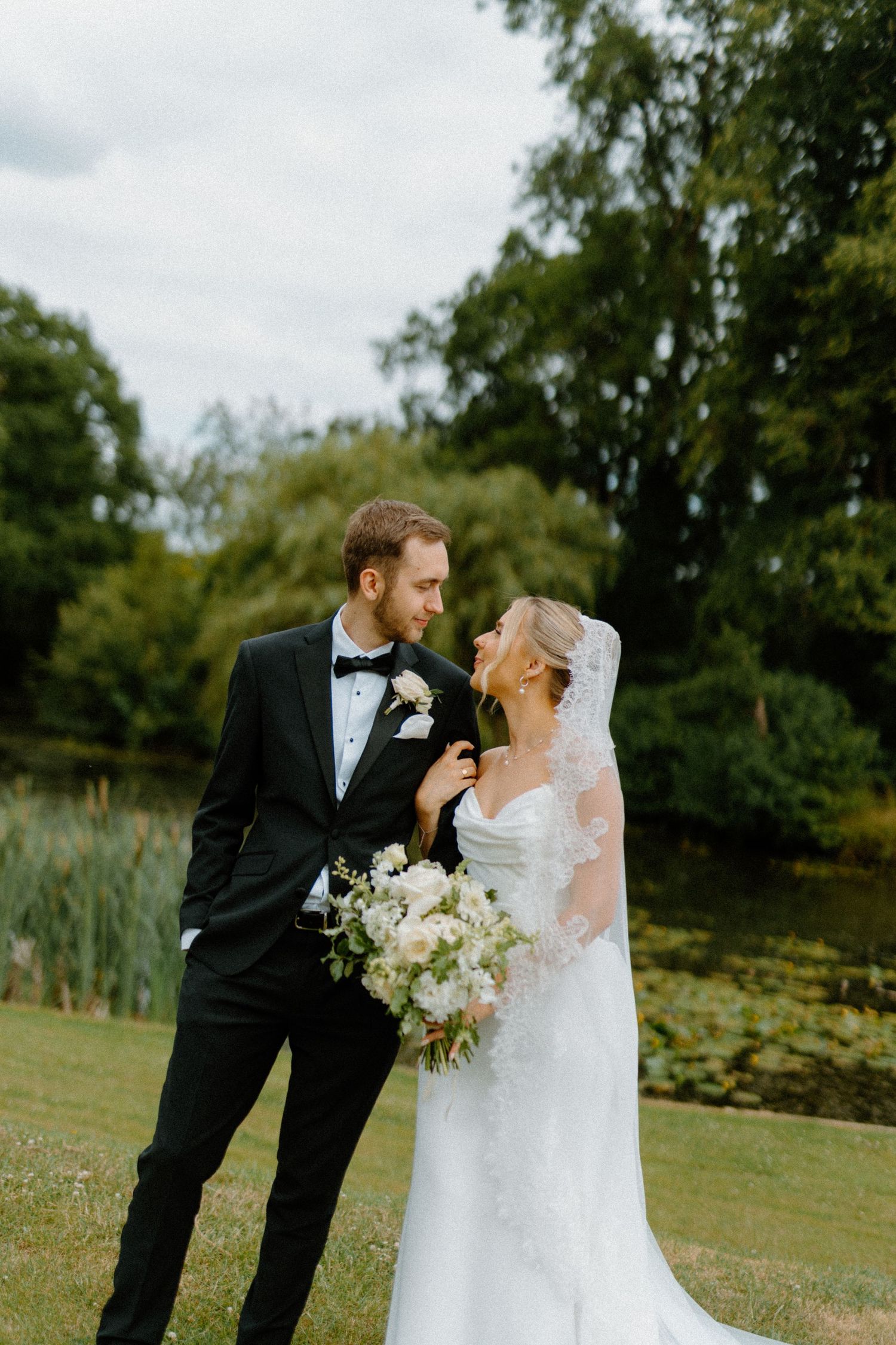 Bride and groom share tender moment near serene pond surrounded by lush green trees on wedding day.