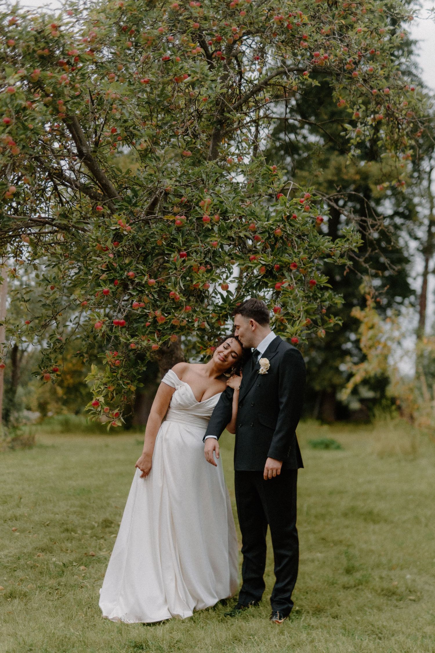Bride and groom share tender moment under red berry tree in lush garden wedding portrait.