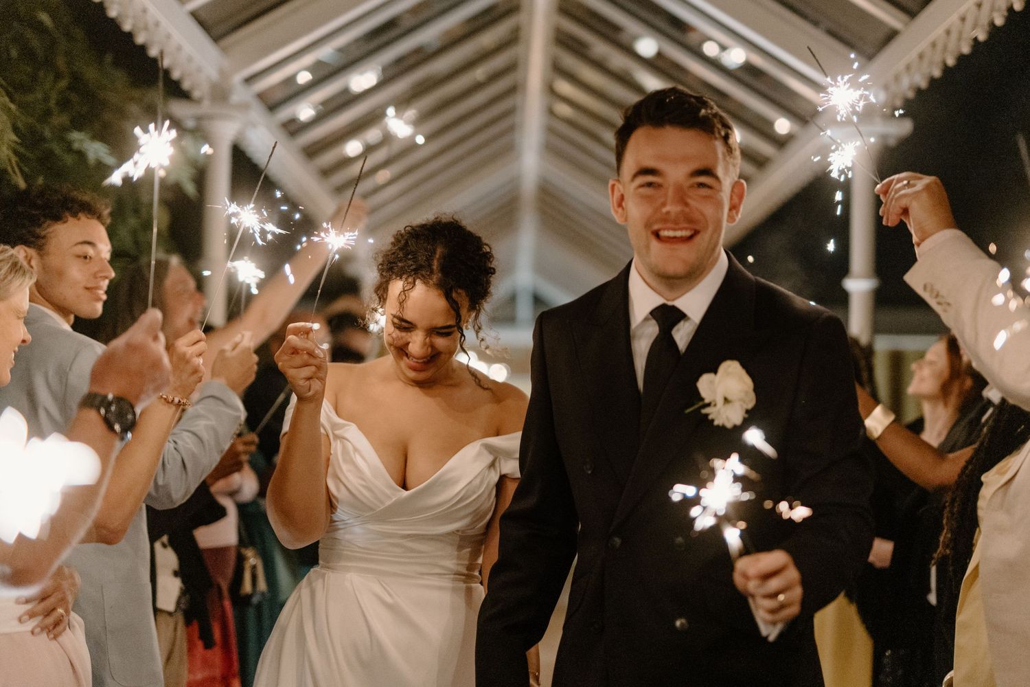 Newlyweds celebrate with sparklers surrounded by cheering guests at night.