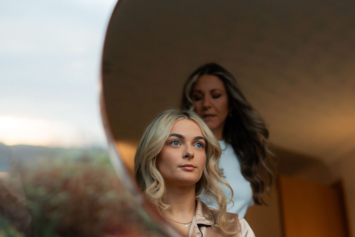 Bride getting hair styled, reflected in round mirror with scenic outdoor backdrop at golden hour.