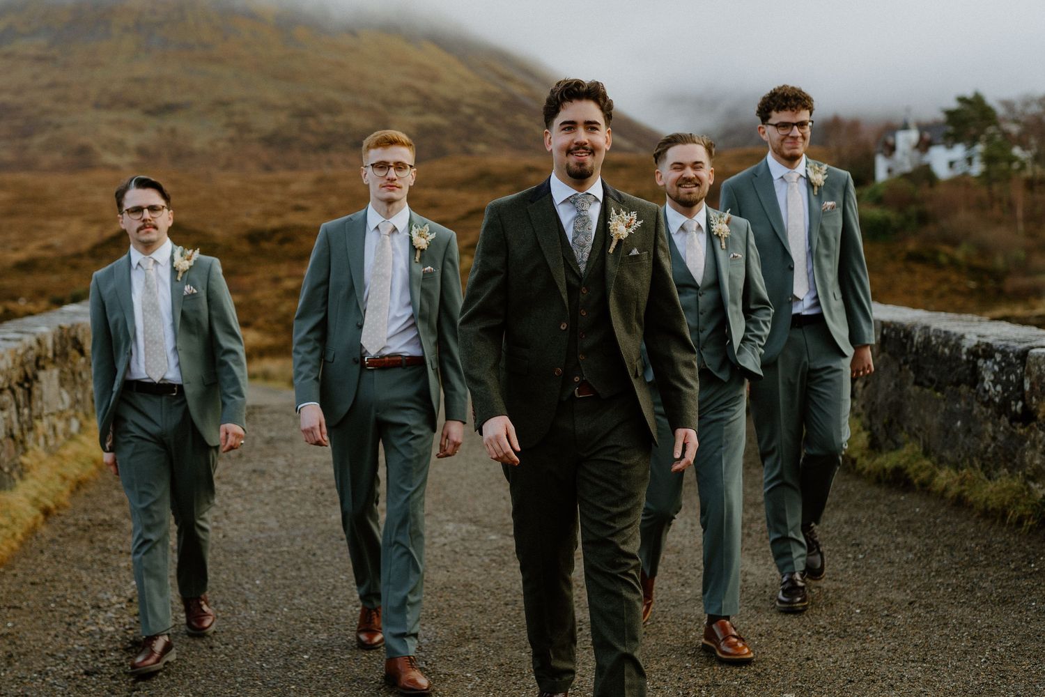 Groom in green tweed suit leads groomsmen in sage suits on misty Scottish highland road.