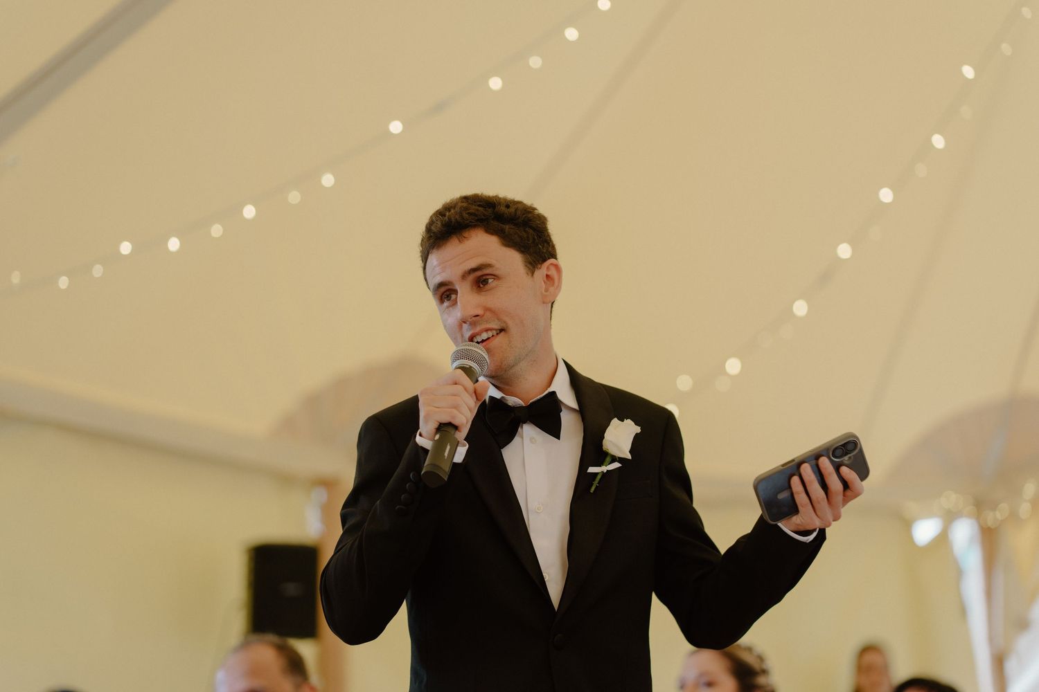Groom in black tuxedo delivering a speech with microphone at elegant wedding reception in marquee.