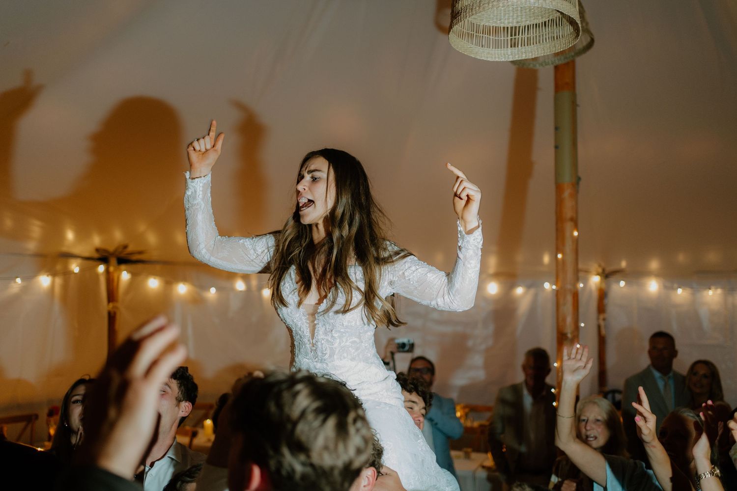 Bride lifted by guests on dance floor at wedding reception under tent with string lights.