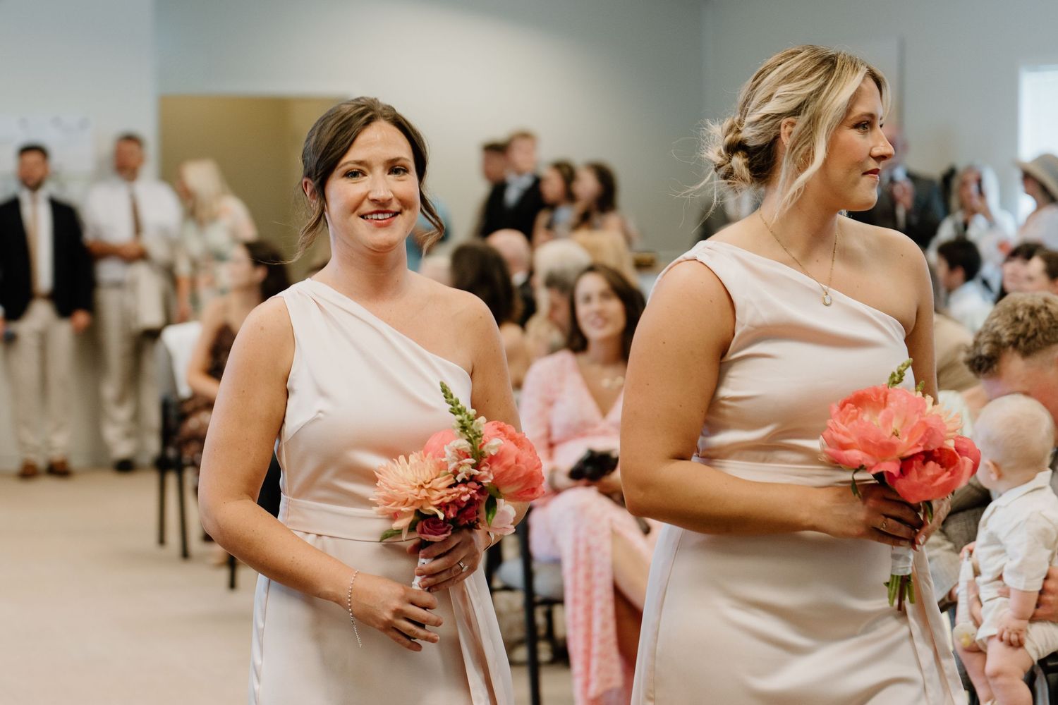 Two bridesmaids in blush one-shoulder dresses carrying coral flowers walking down wedding aisle.