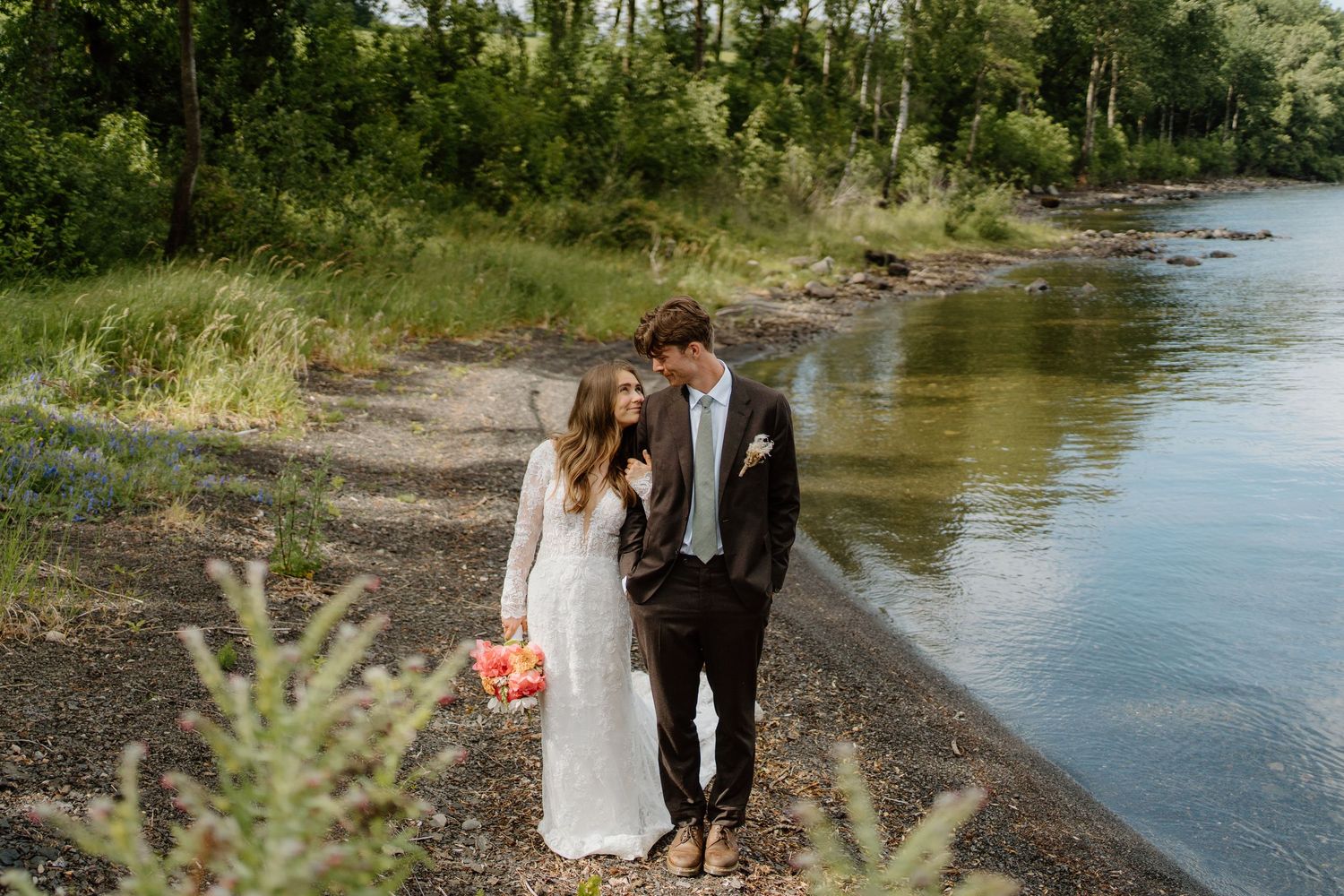 Bride and groom walking hand in hand along a scenic rocky riverbank surrounded by lush greenery.