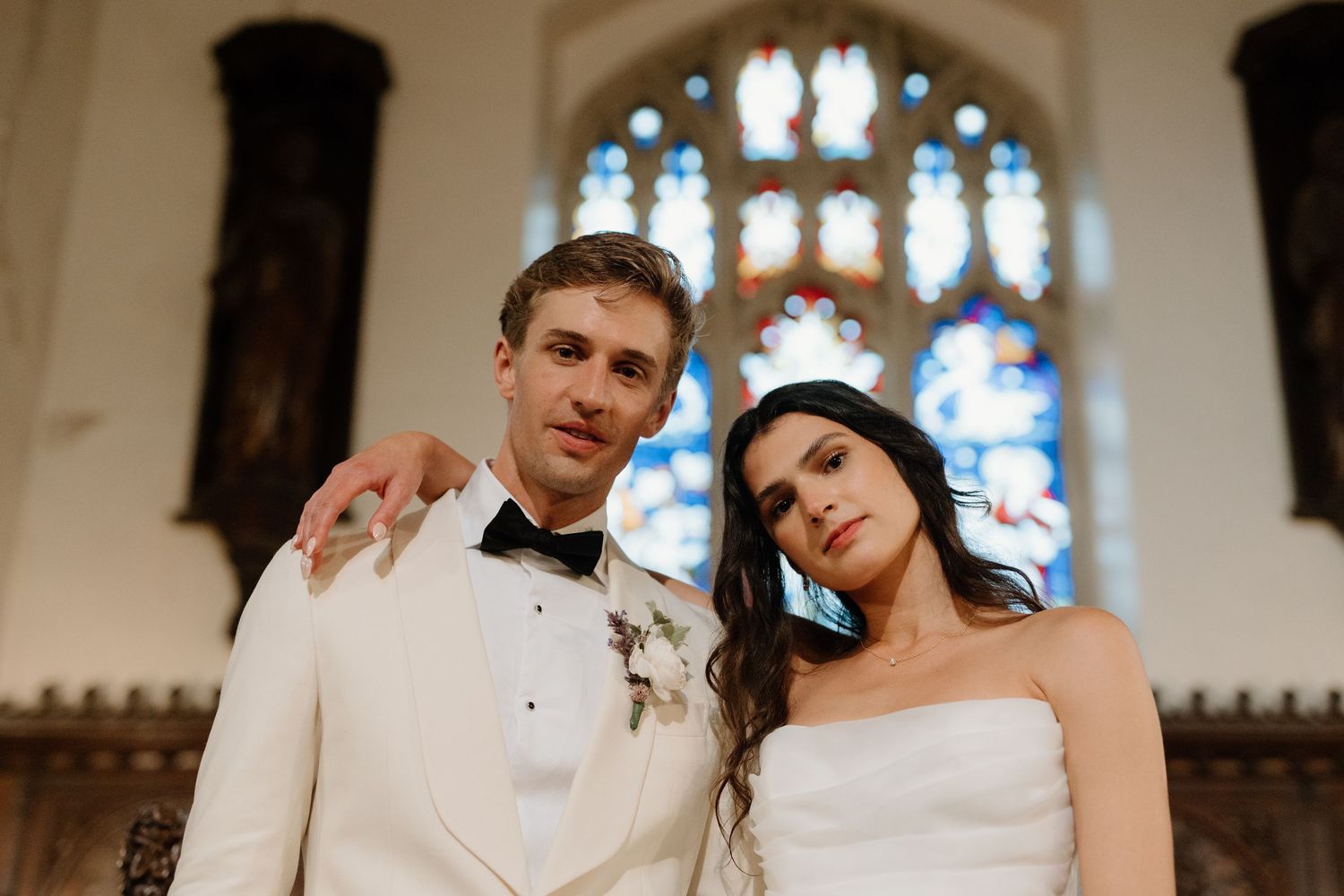 Bride and groom pose solemnly inside a church with stunning stained glass windows glowing behind them.