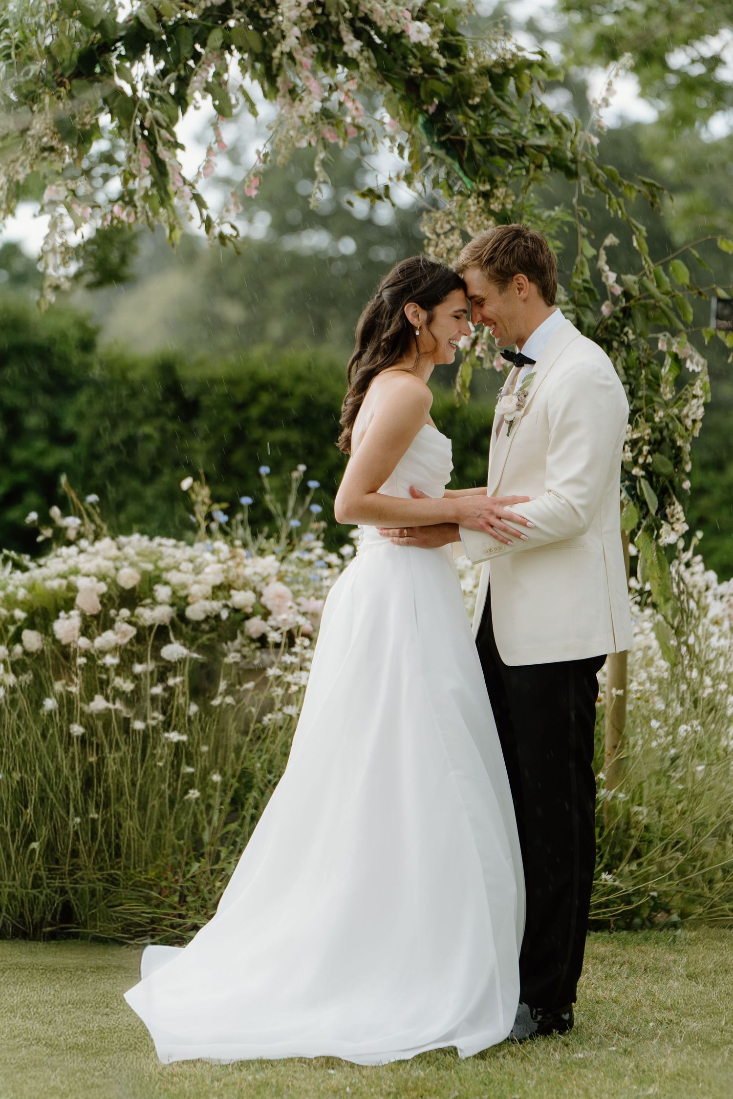 Bride and groom touching foreheads tenderly under floral arch in blooming garden setting.