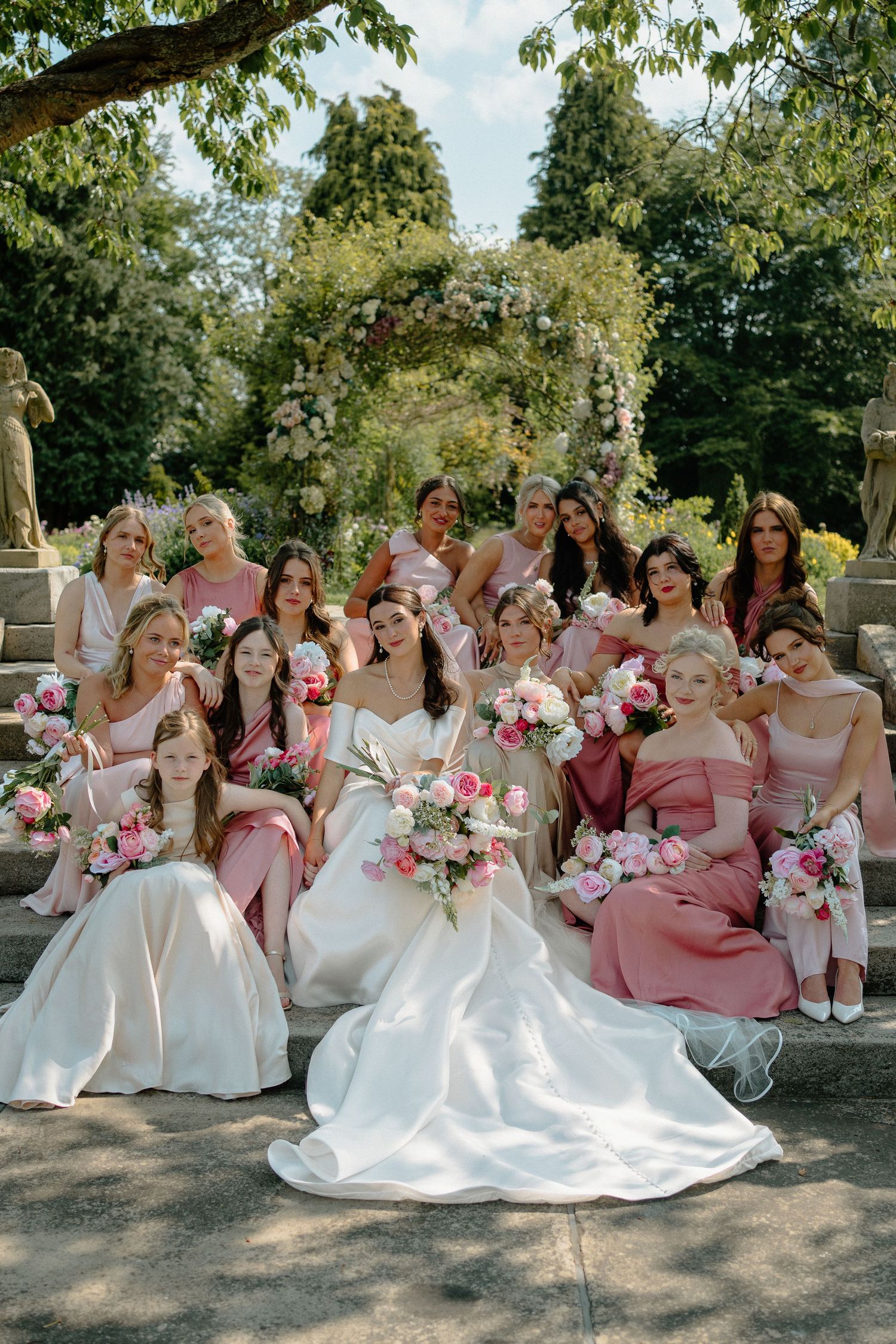 Bride in white gown poses with bridesmaids in pink dresses holding floral bouquets on garden steps.