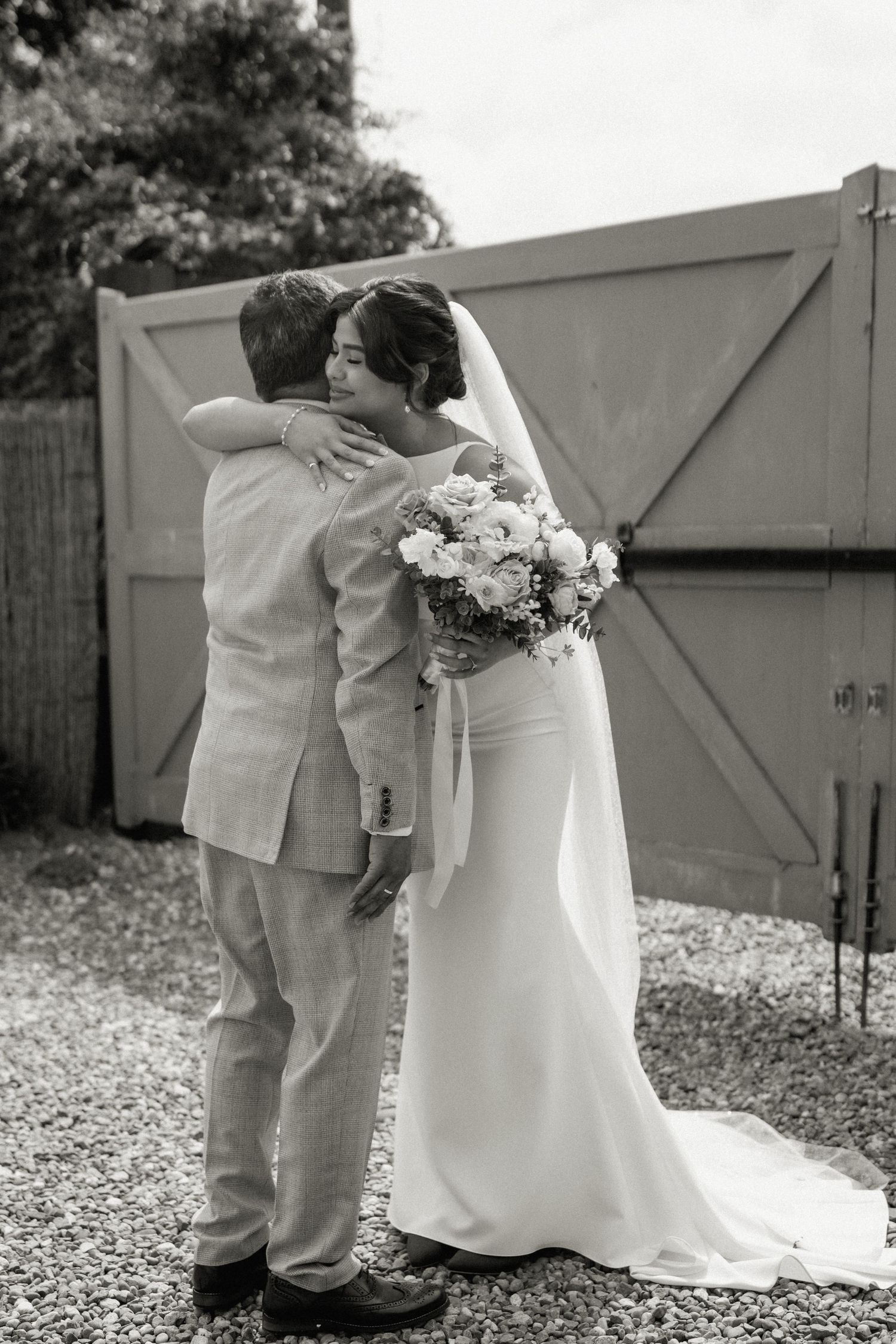 Black and white photo of bride and groom embracing tenderly in front of large wooden gates.