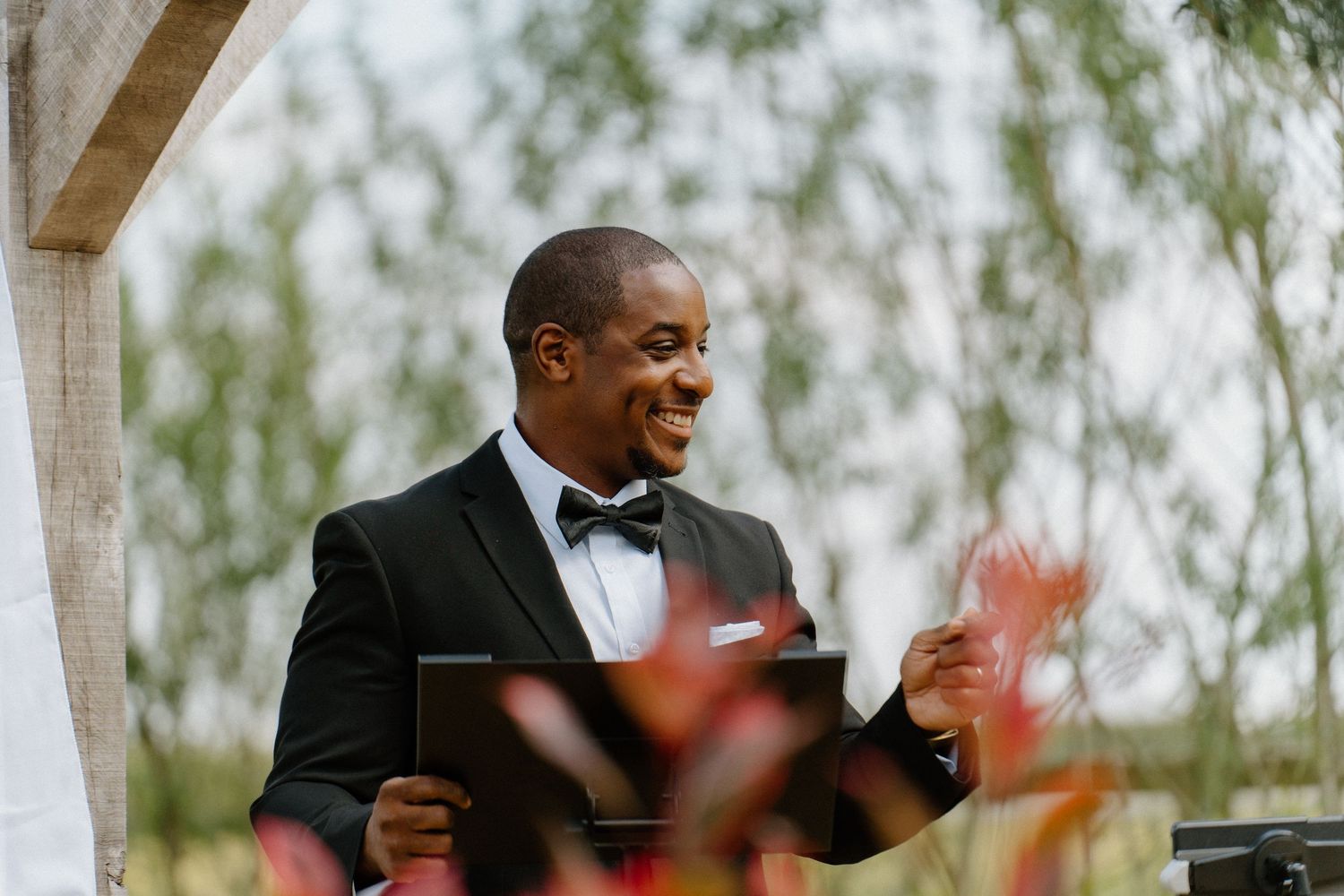 Smiling officiant in black tuxedo holds ceremony booklet while standing beneath wooden arch with greenery.