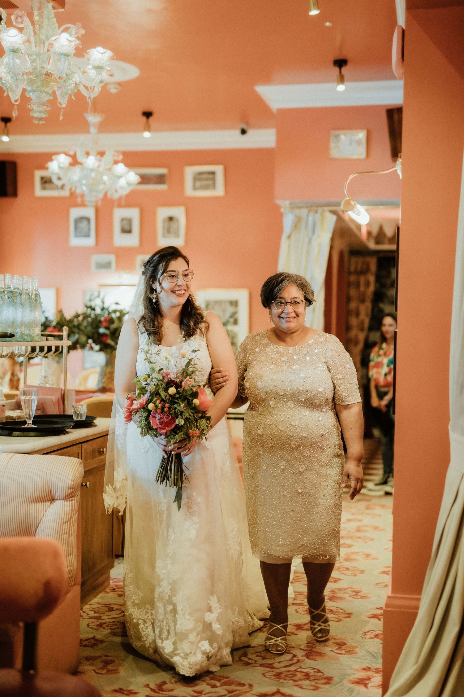 Smiling bride in lace gown holding bouquet walks with older woman in elegant venue.