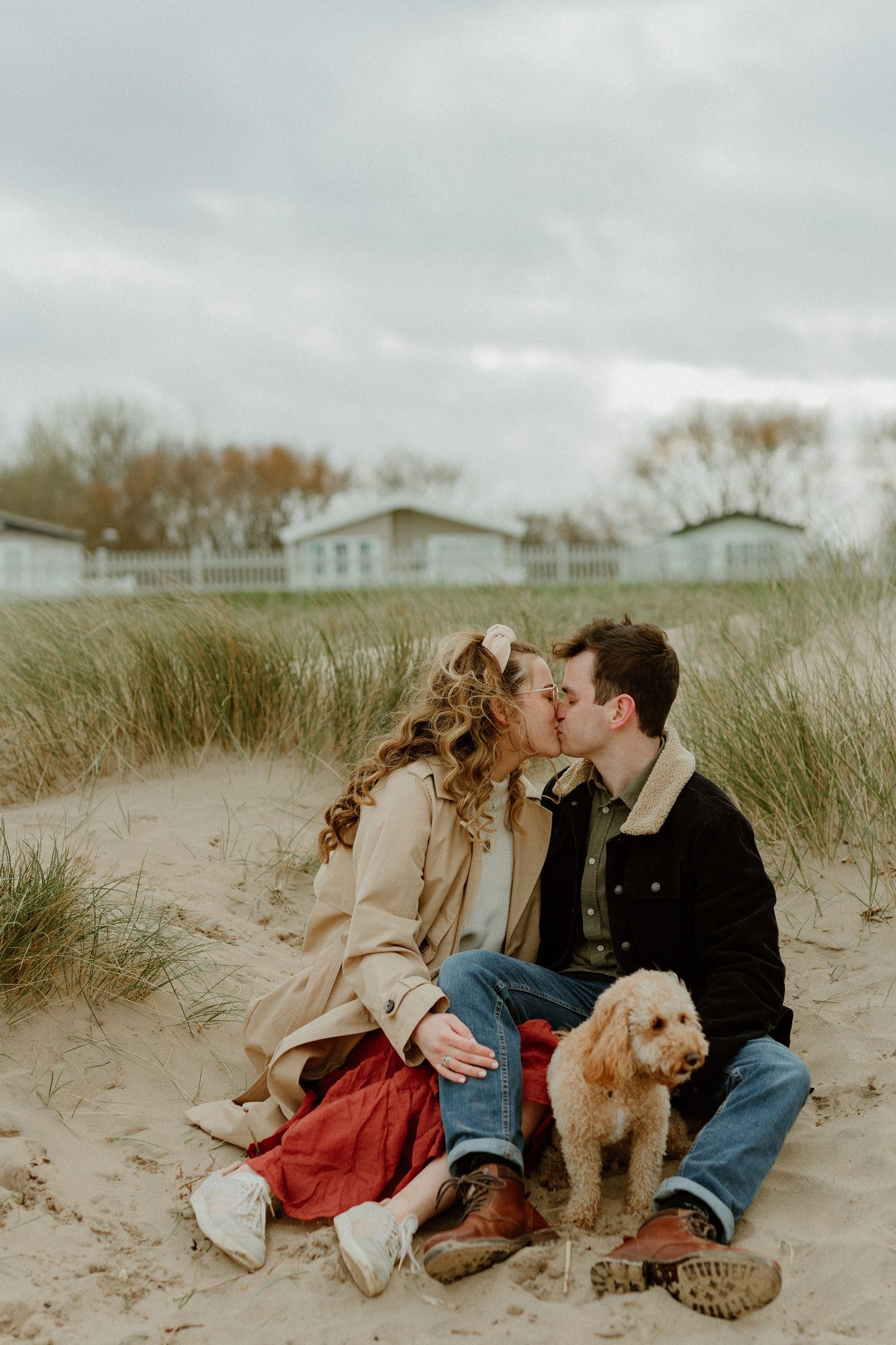 Couple kissing in sand dunes with an adorable golden doodle dog beside them.