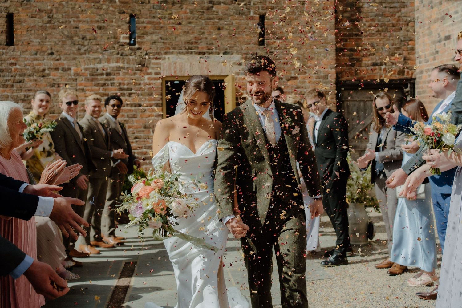 Bride and groom walk through confetti shower outside rustic brick barn venue surrounded by smiling wedding guests.