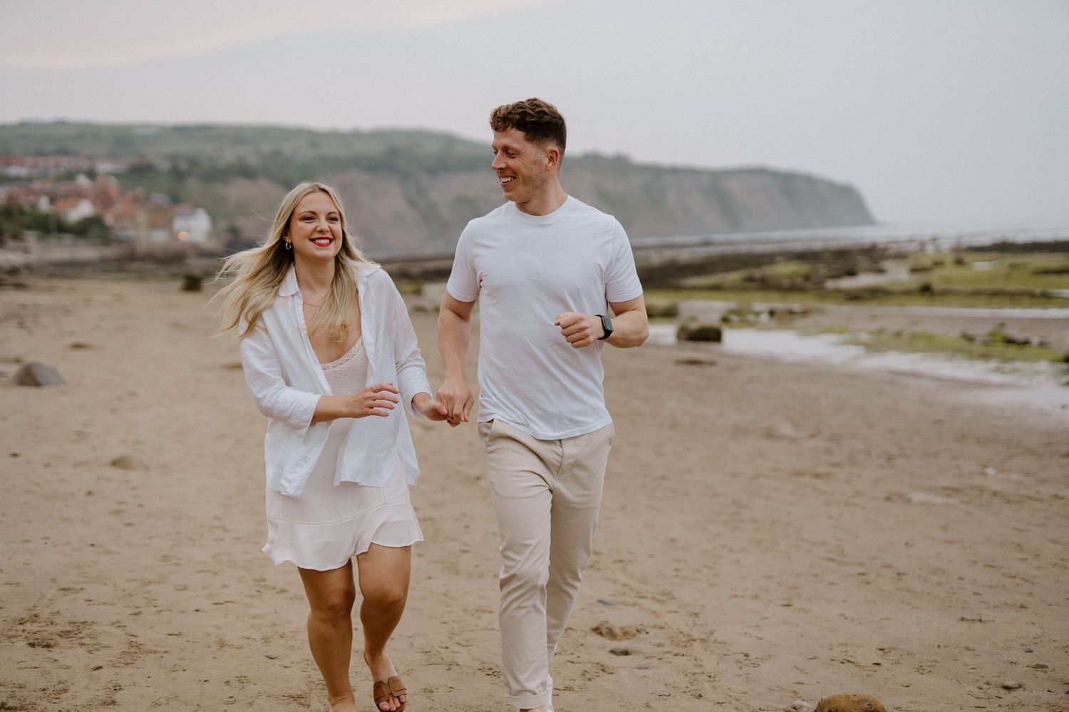 Happy couple laughing and running hand in hand along a sandy beach with coastal cliffs behind.