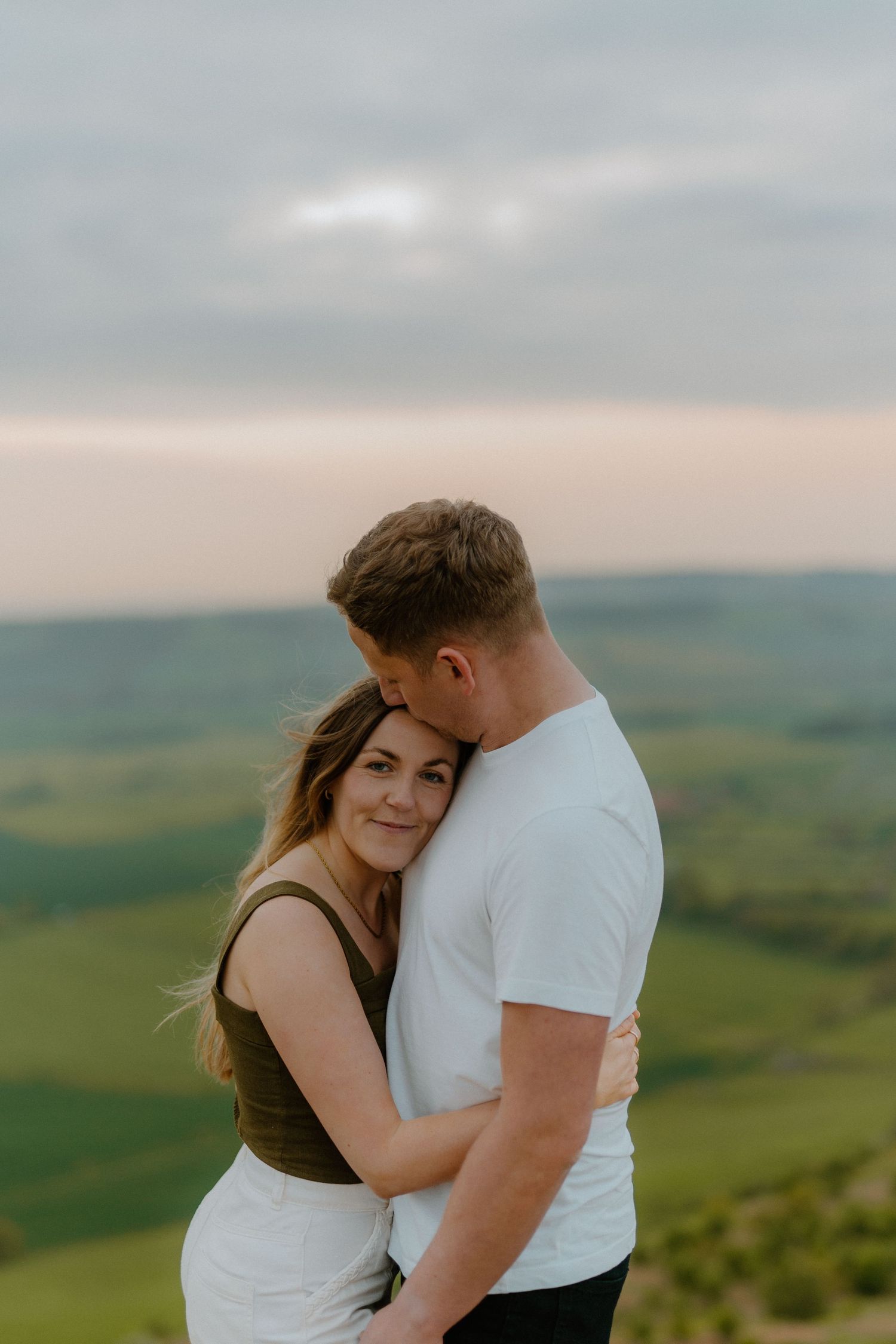 Couple embracing at sunset with expansive green countryside landscape behind them.