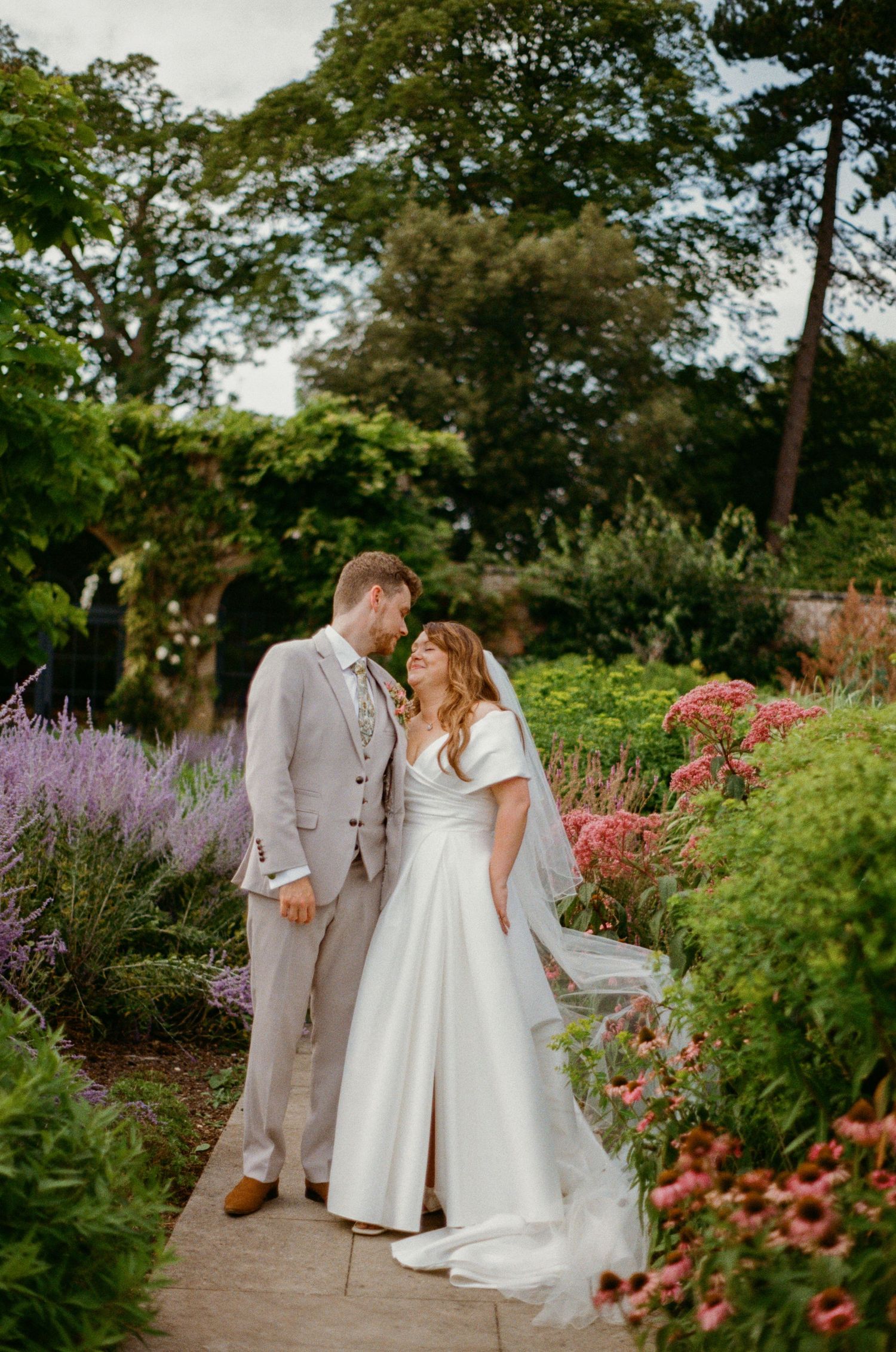 Bride and groom share romantic moment on garden path surrounded by vibrant lavender and wildflowers in bloom.