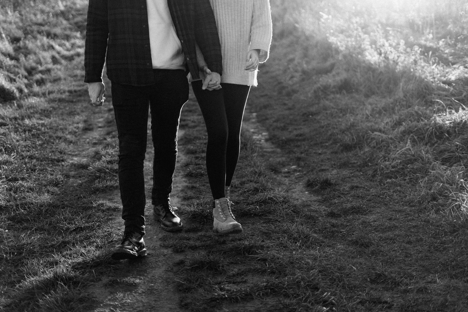 Black and white photo of couple holding hands walking on grass path, romantic outdoor shoot.