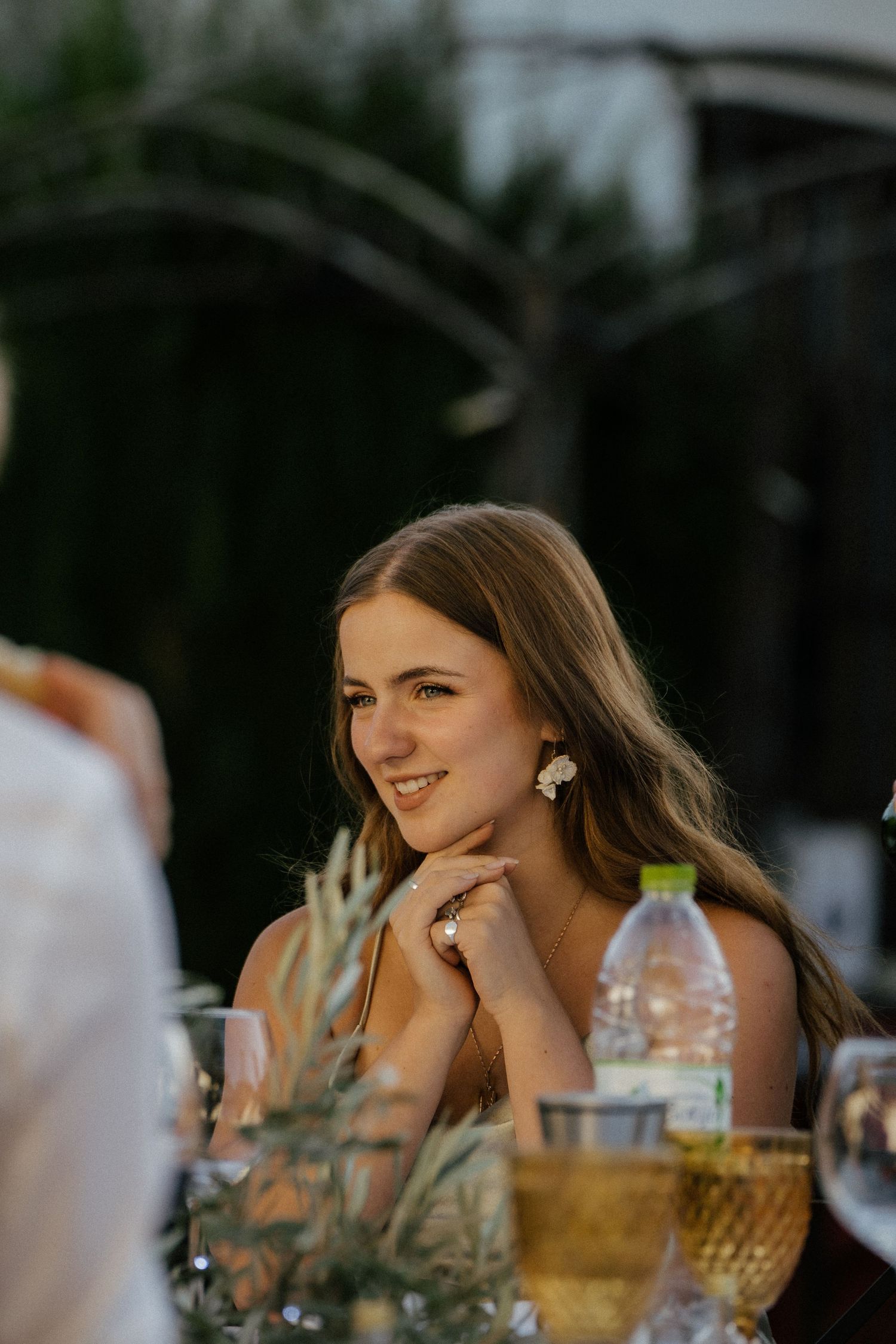 Smiling wedding guest with floral earrings seated at outdoor reception table at golden hour.