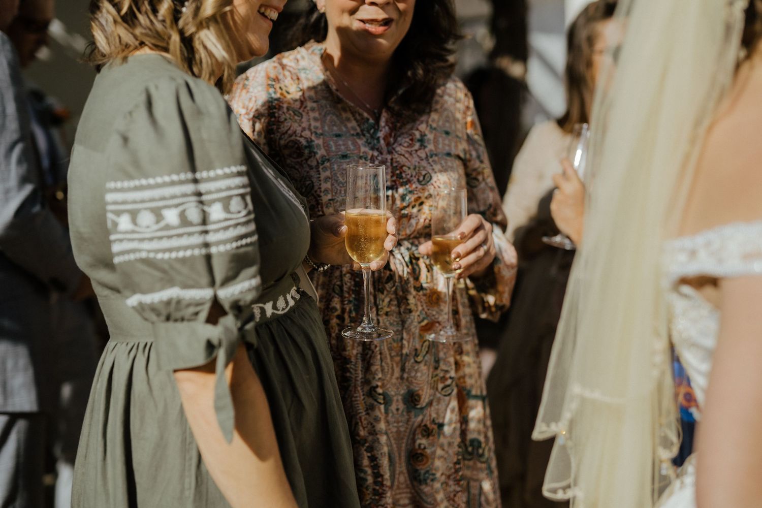 Wedding guests in elegant dresses holding champagne glasses at outdoor reception.