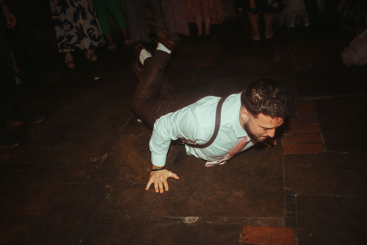 A man in suspenders and dress shirt crawling on a dark dance floor at a wedding reception.