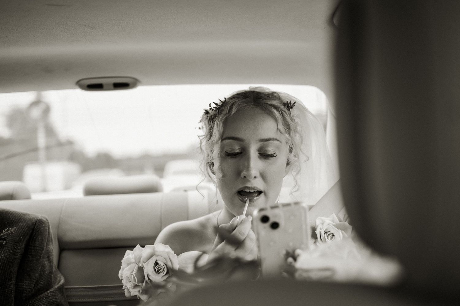 Black and white photo of bride touching up makeup using phone mirror inside wedding car.