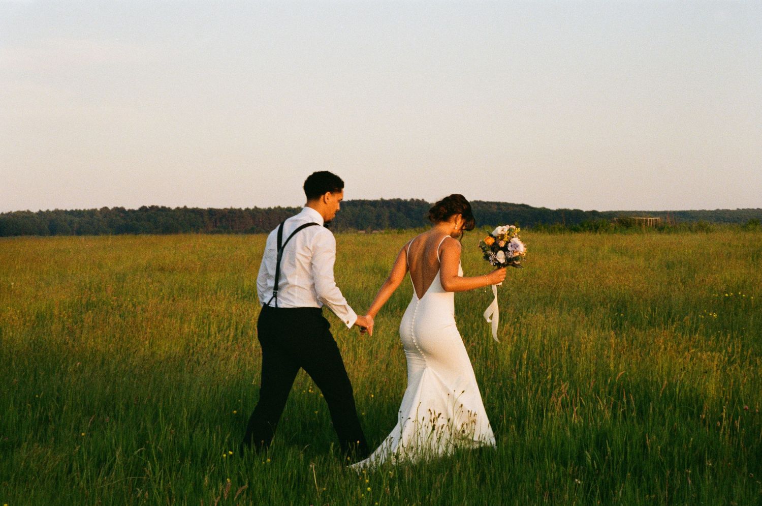 Bride and groom walking hand in hand through lush green field at golden hour sunset.