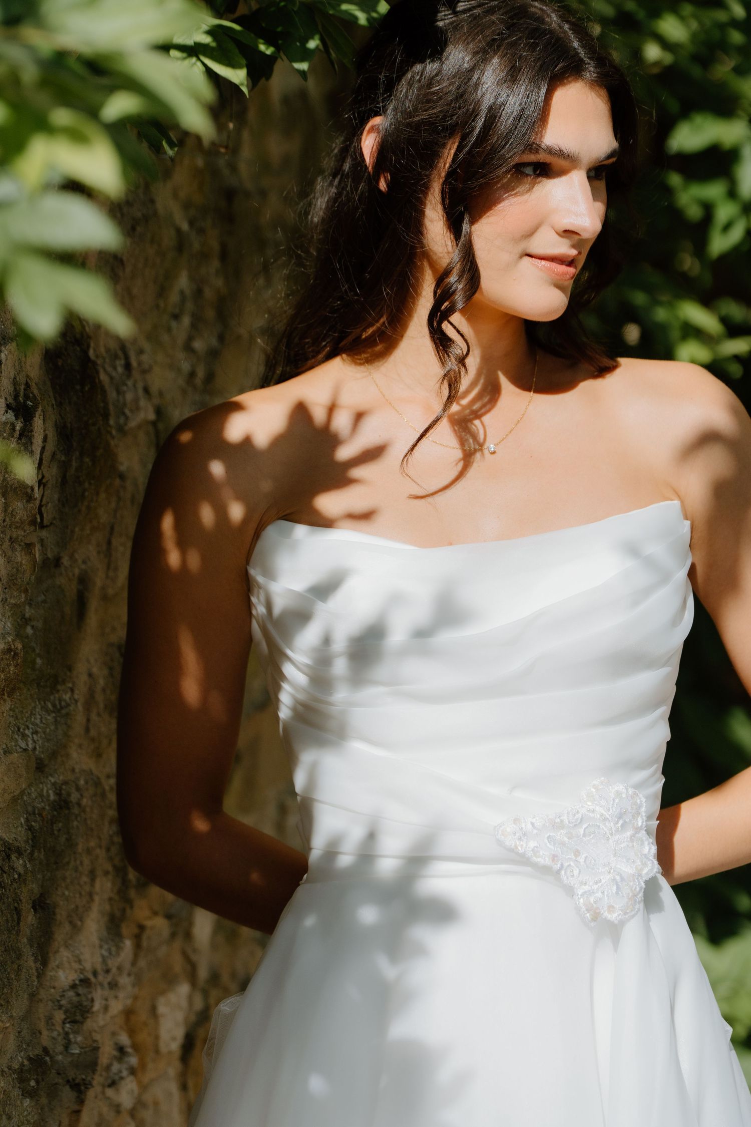 Elegant bride in strapless white gown leans against a tree in dappled sunlight outdoors.