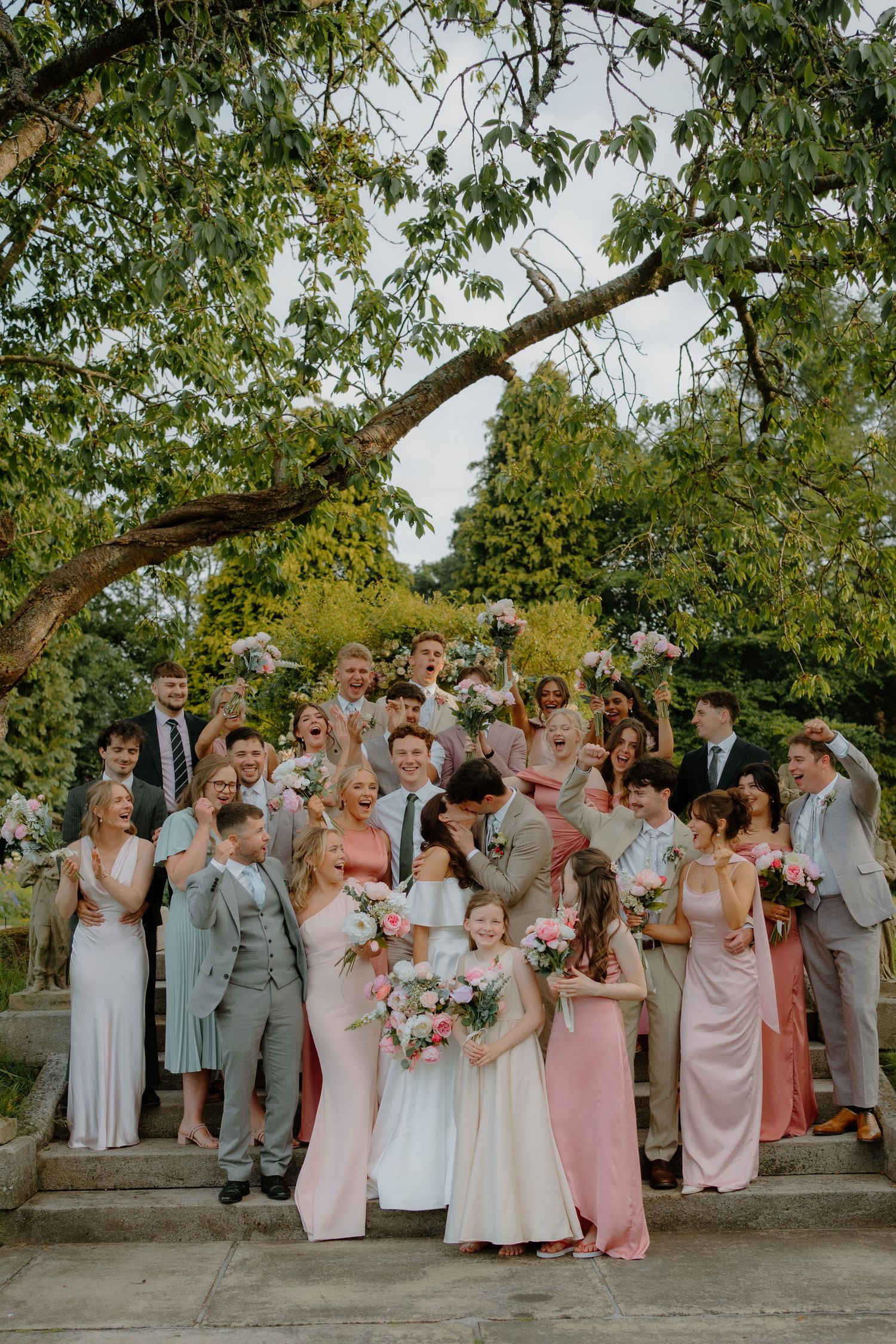 Large wedding party group photo on outdoor steps with bride and groom kissing in center.