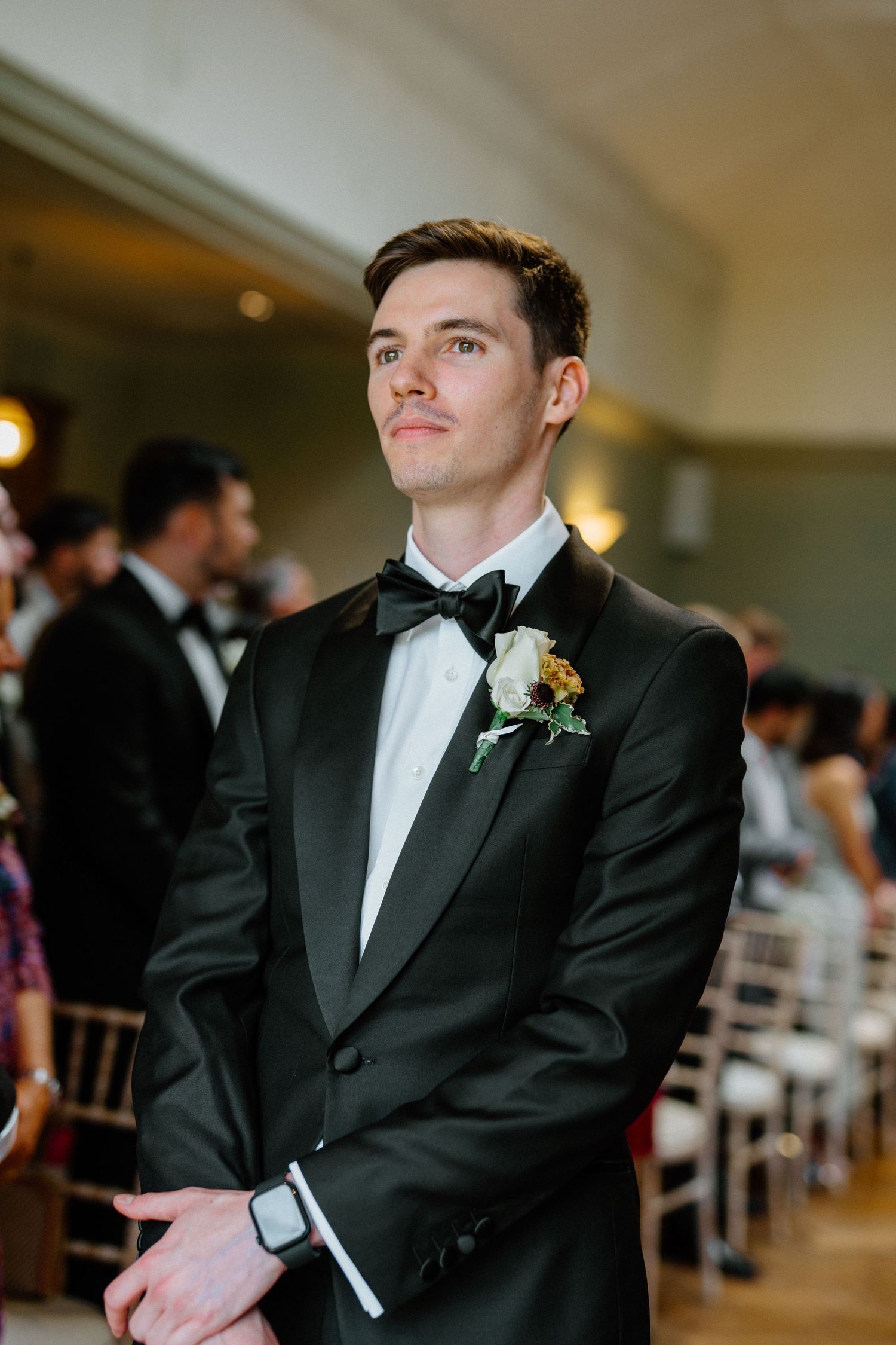 Groom in black tuxedo with floral boutonniere waiting at altar during wedding ceremony.