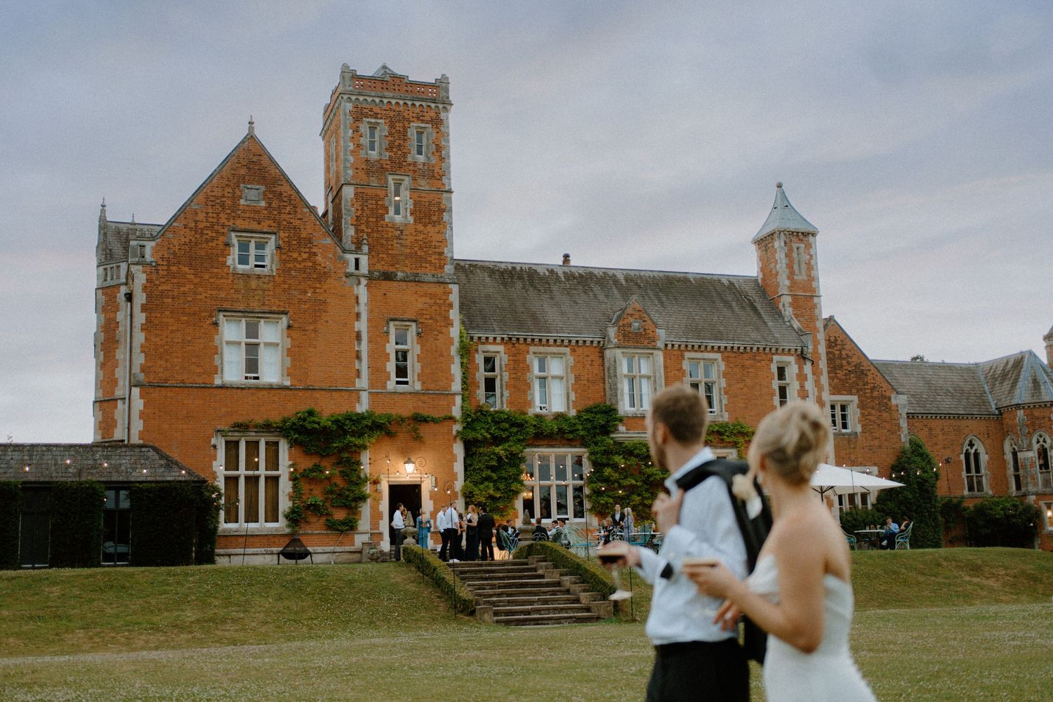 Bride and groom walking across lawn toward a grand red-brick English manor house wedding venue on a cloudy day.