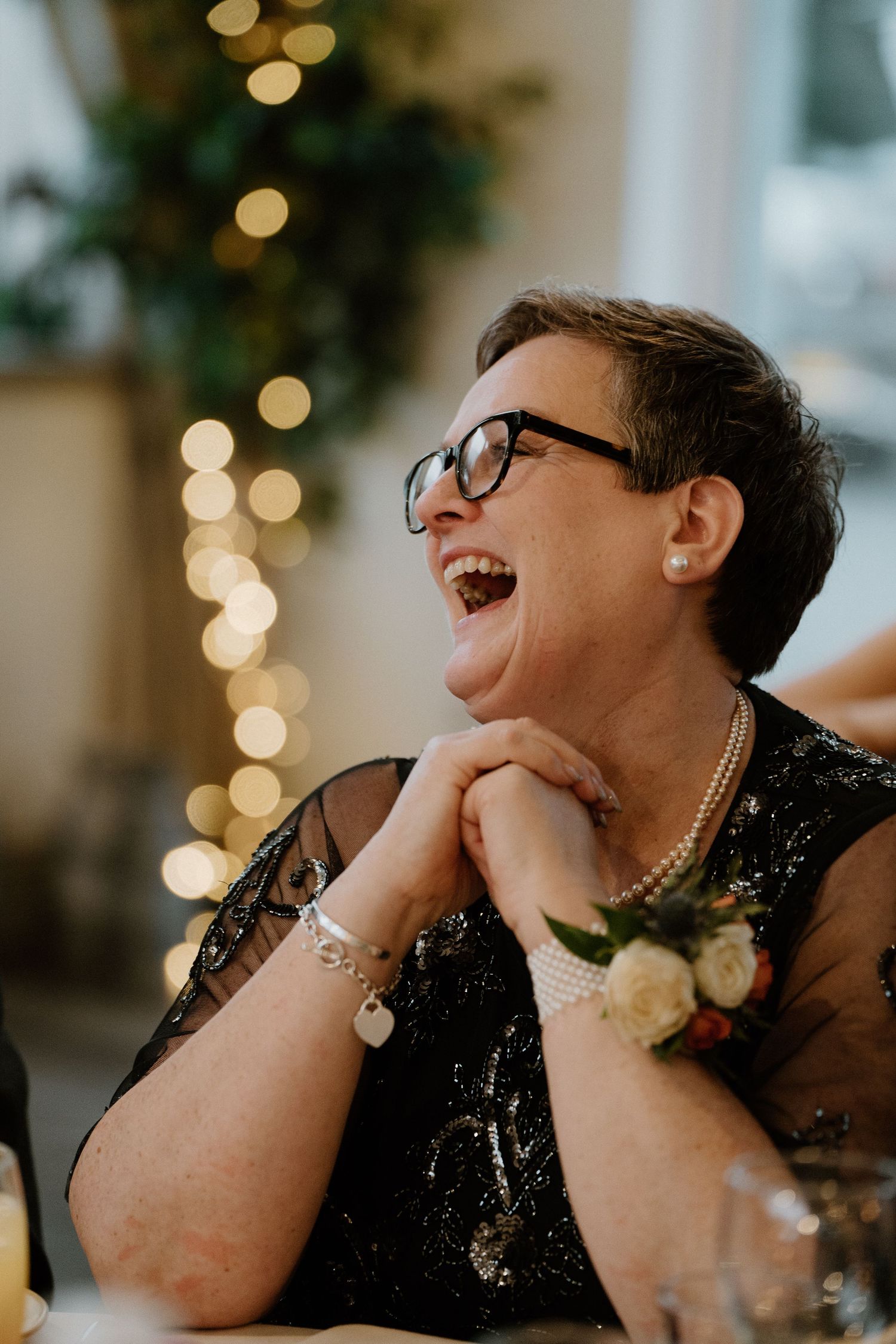 Joyful older woman in black sequin dress laughing at a wedding reception with bokeh lights behind her.