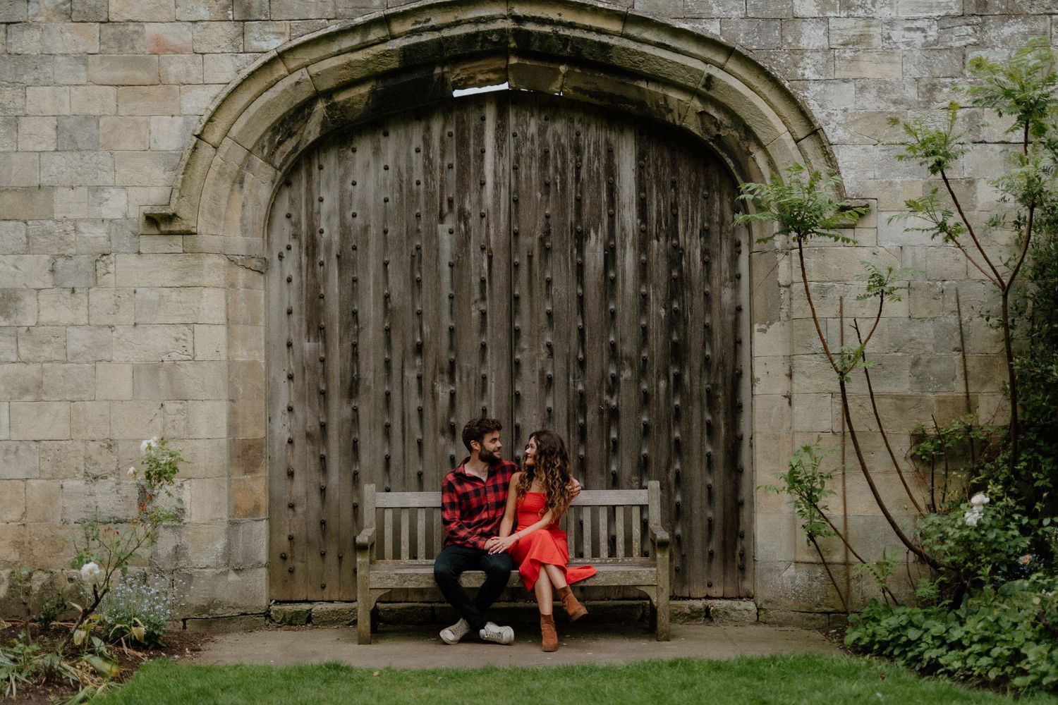 Couple sitting on a wooden bench in front of a rustic stone arched wall in a garden.
