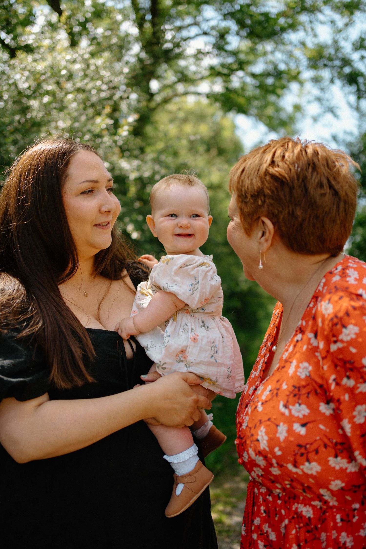 Young mother in black dress holding smiling baby girl while older woman in red looks on outdoors.