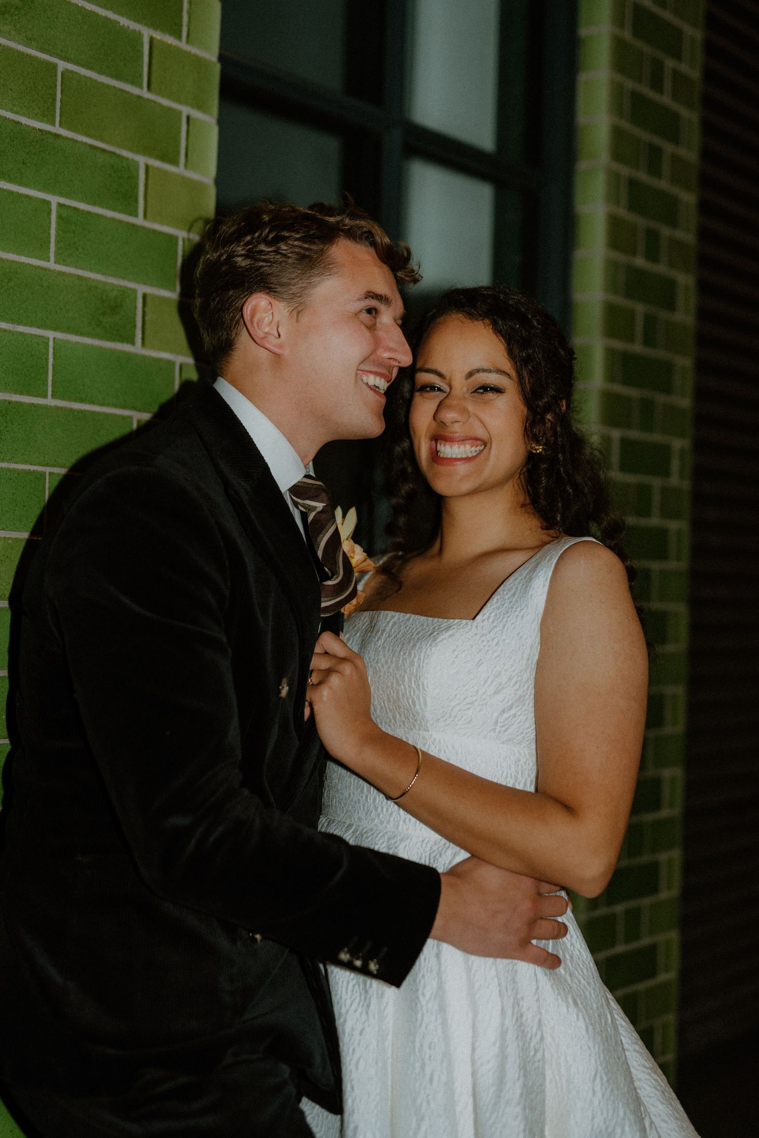 A smiling bride in a white dress embraces her groom in a black velvet suit against a green brick wall.