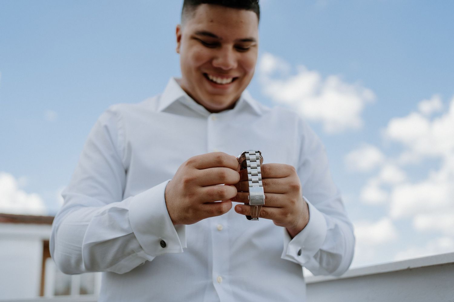 Smiling groom in white shirt holding wedding watch under blue sky before his wedding ceremony.