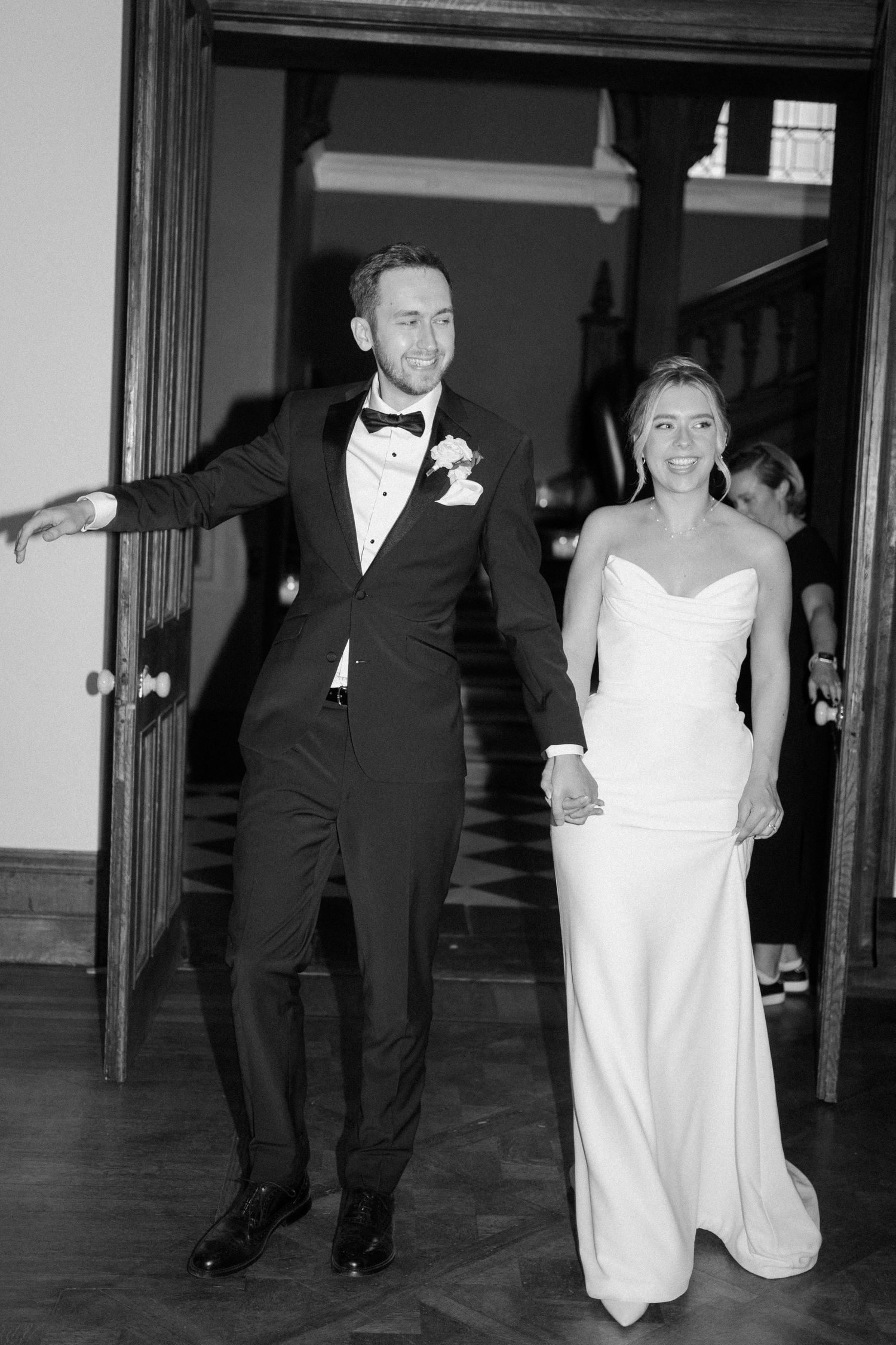 Black and white photo of smiling bride and groom holding hands making grand entrance at elegant wedding reception.