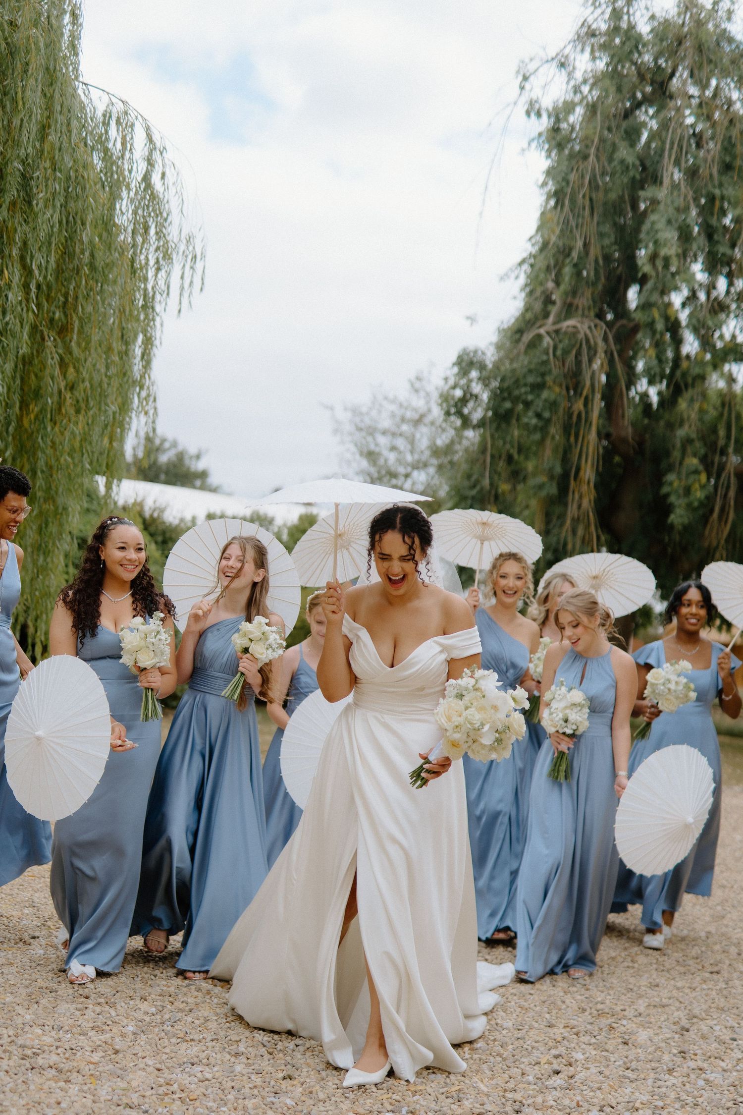 Radiant bride in white gown leading bridesmaids in blue dresses carrying white parasols.