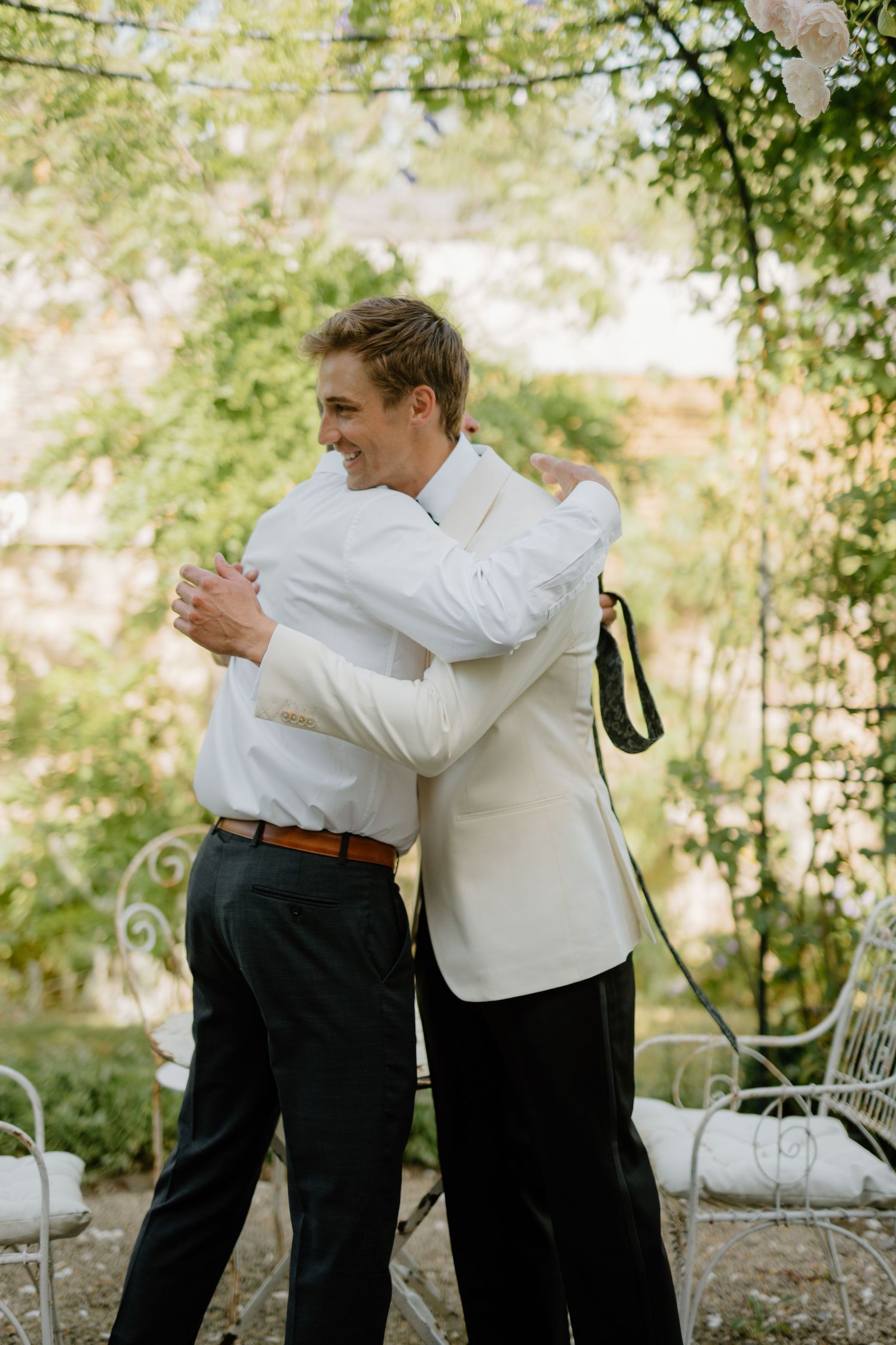 Two grooms share a heartfelt hug during their outdoor wedding ceremony surrounded by lush green trees.