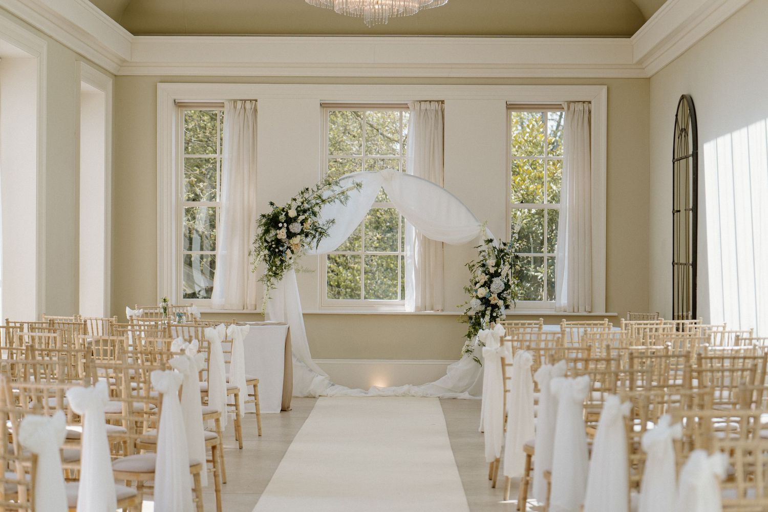 Elegant wedding ceremony room with white chairs, floral arch, and natural light streaming through tall windows.